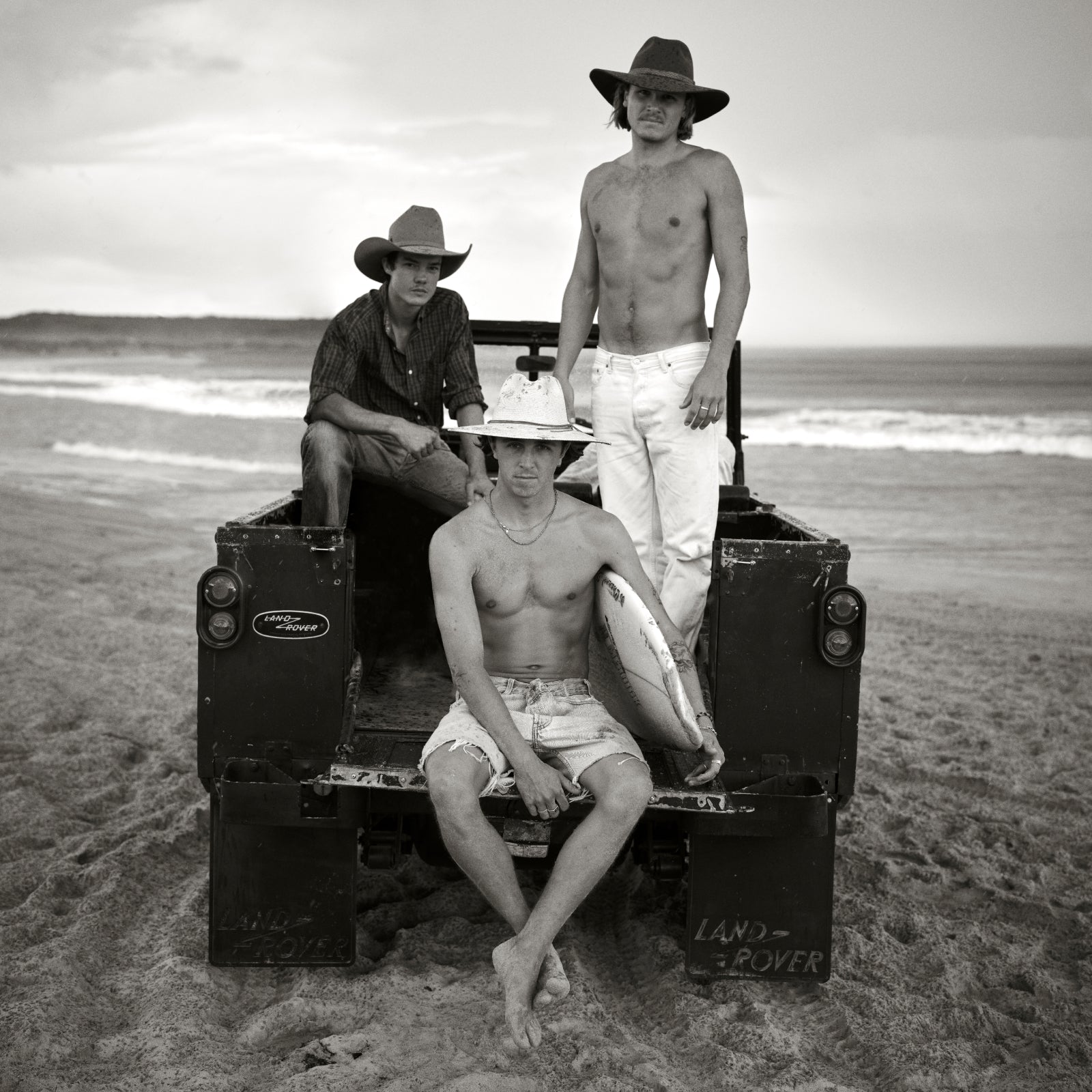 Three men on a beach with a Land Rover, one holding a surfboard, wearing Akubra hats
