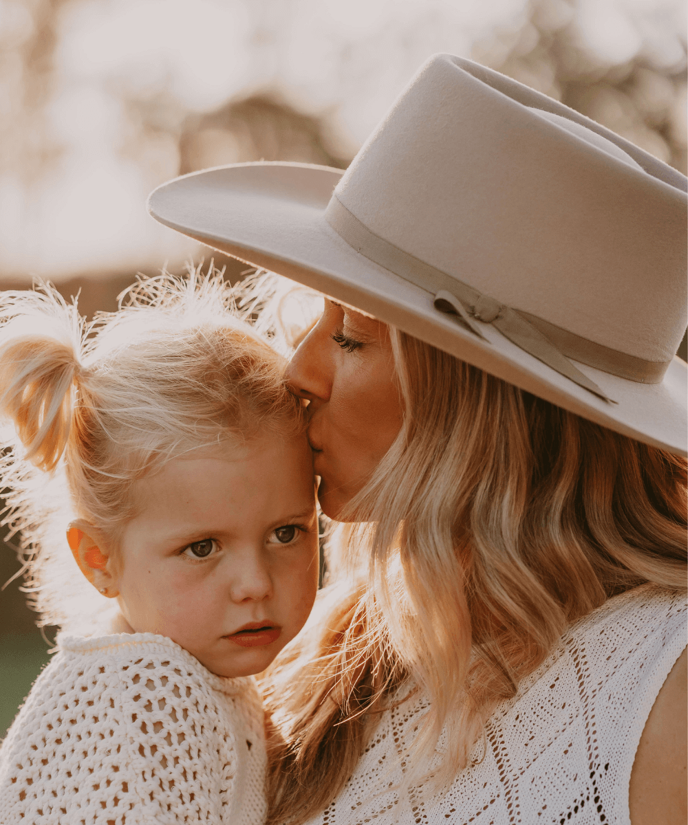 Wopman holding a child and kissing them on their forehead. The woman is wearing Akubra Warrego hat in sand colour.