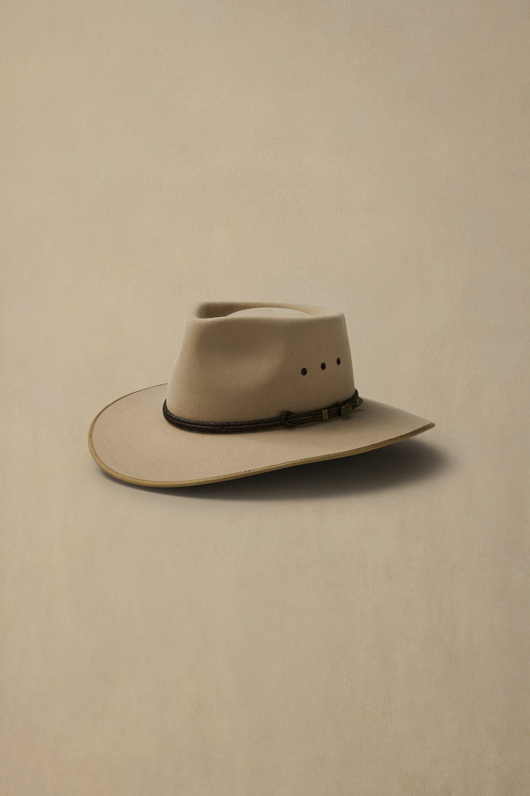 Sand-colored felt cowboy hat with teardrop crown and dark leather hatband on neutral beige background