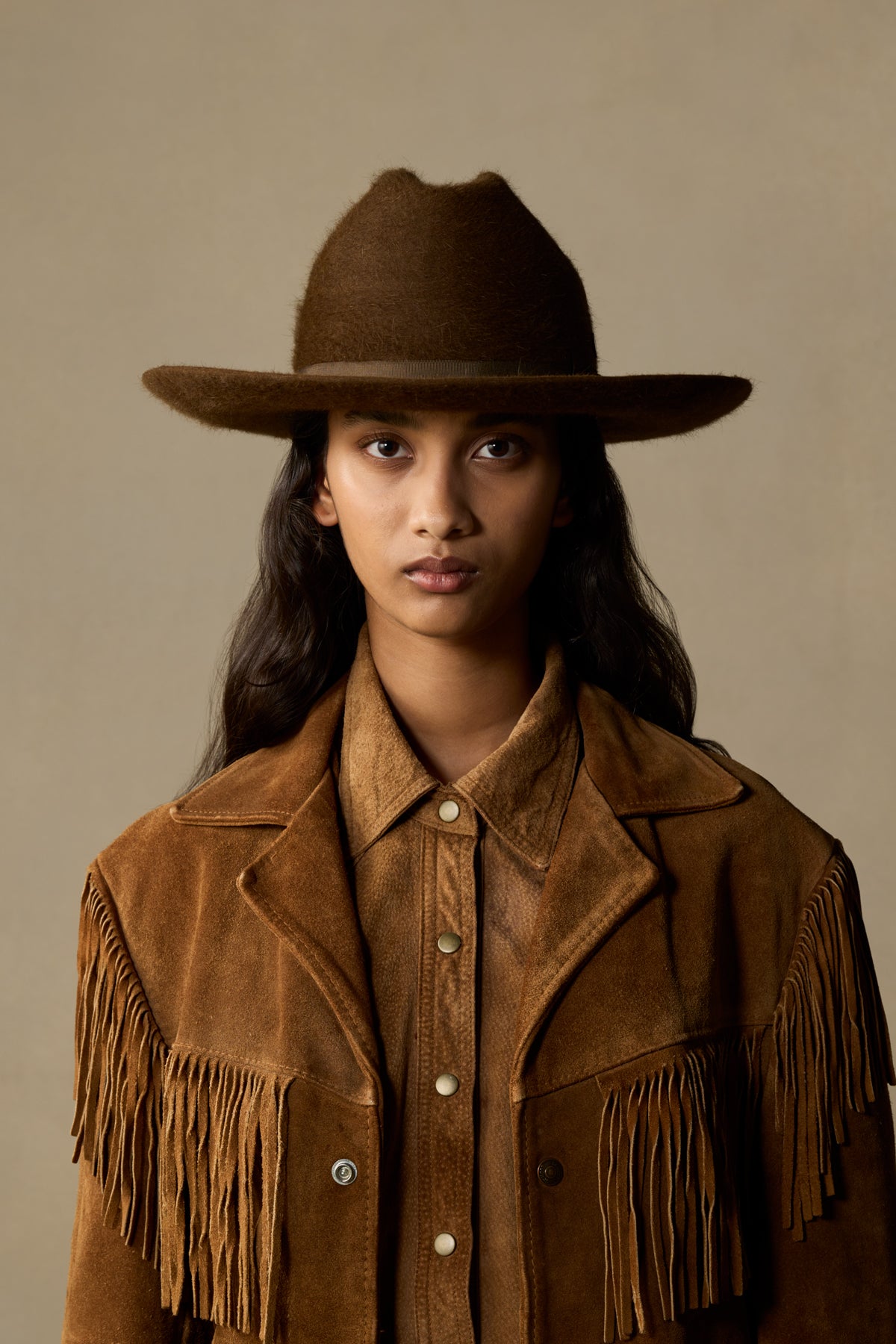 Brown felt cowboy hat with fringed suede jacket on model with long dark hair against neutral background