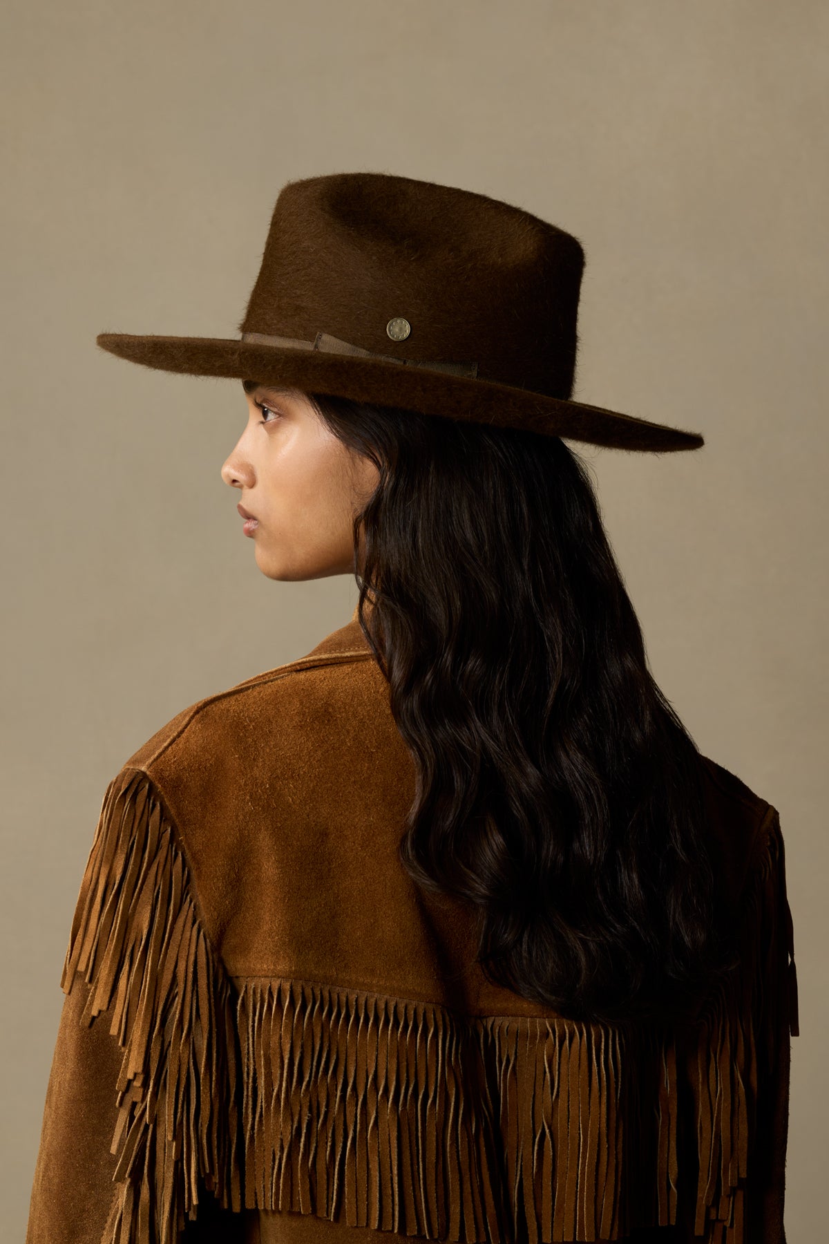 Brown felt cowboy hat with silver badge on woman wearing fringed suede jacket, side profile view