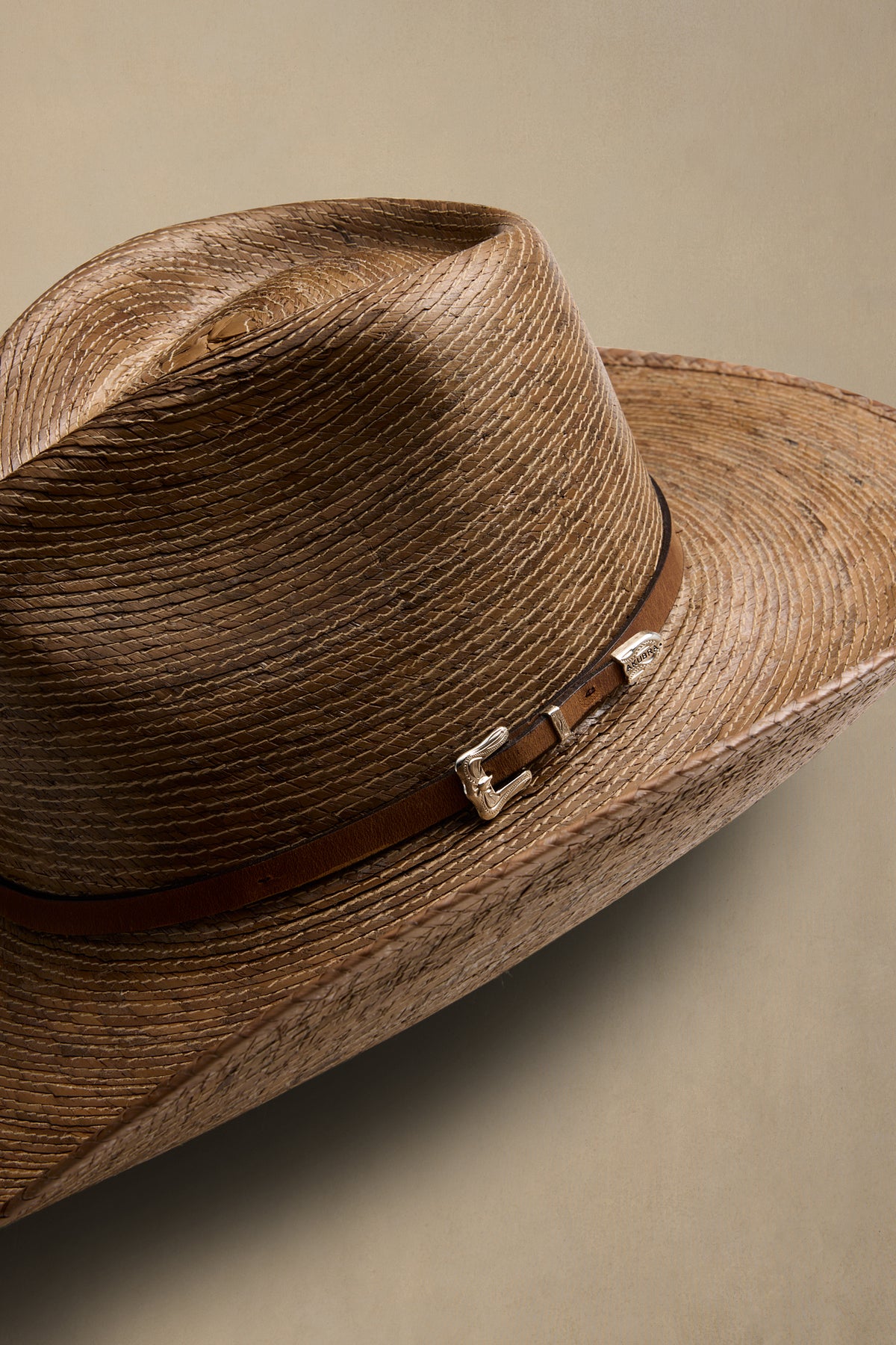 Tan straw cowboy hat with brown leather band and silver buckle detail against beige background