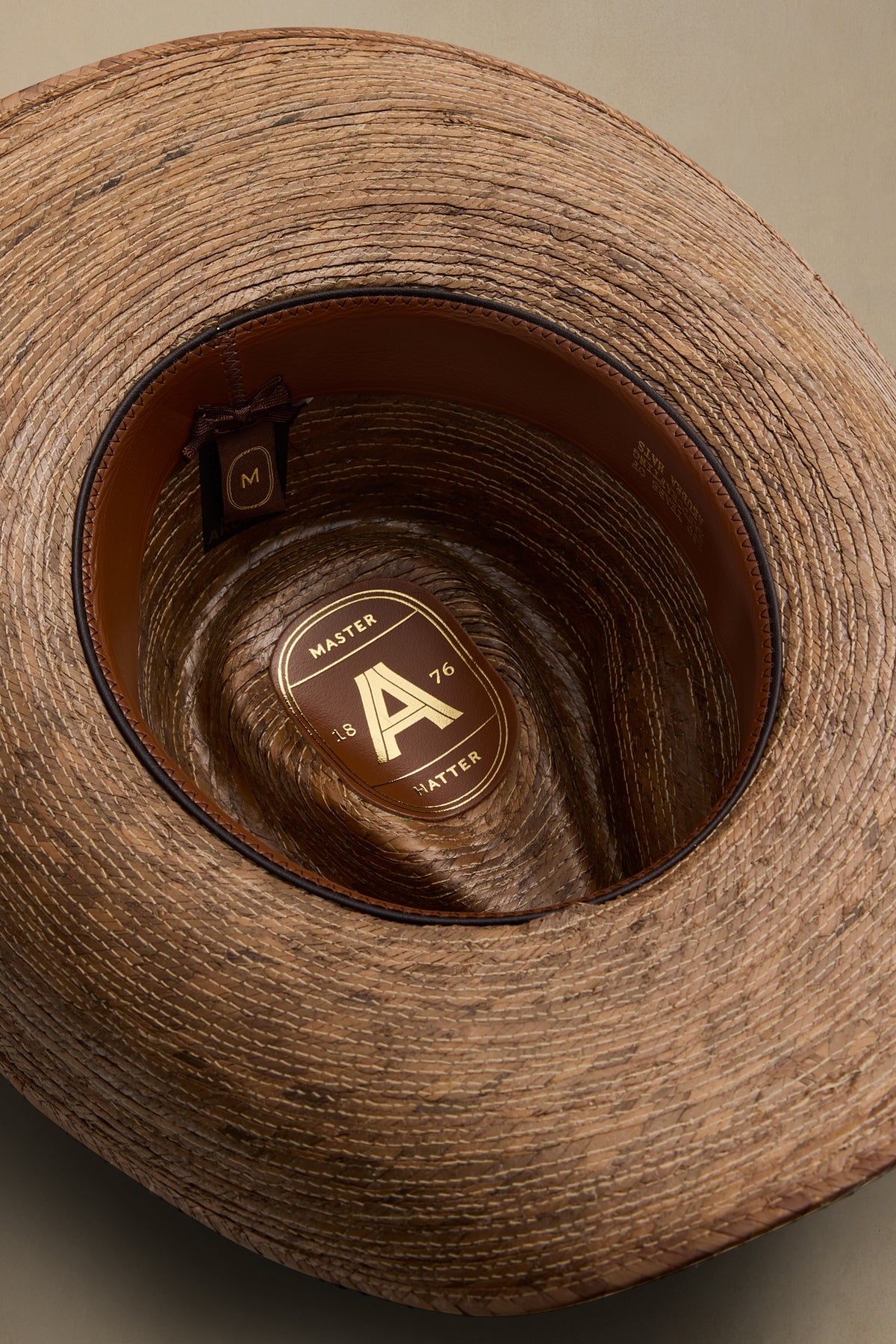 Overhead view of tan straw hat with brown leather band and gold "A" badge showing size and style details