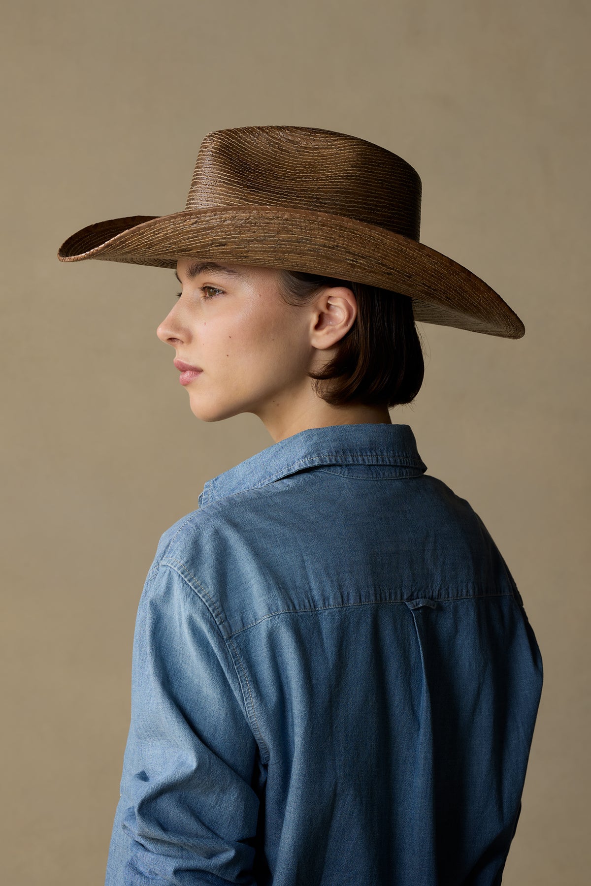 Woman wearing dark tan straw cowboy hat and blue denim shirt, shown in profile against beige background