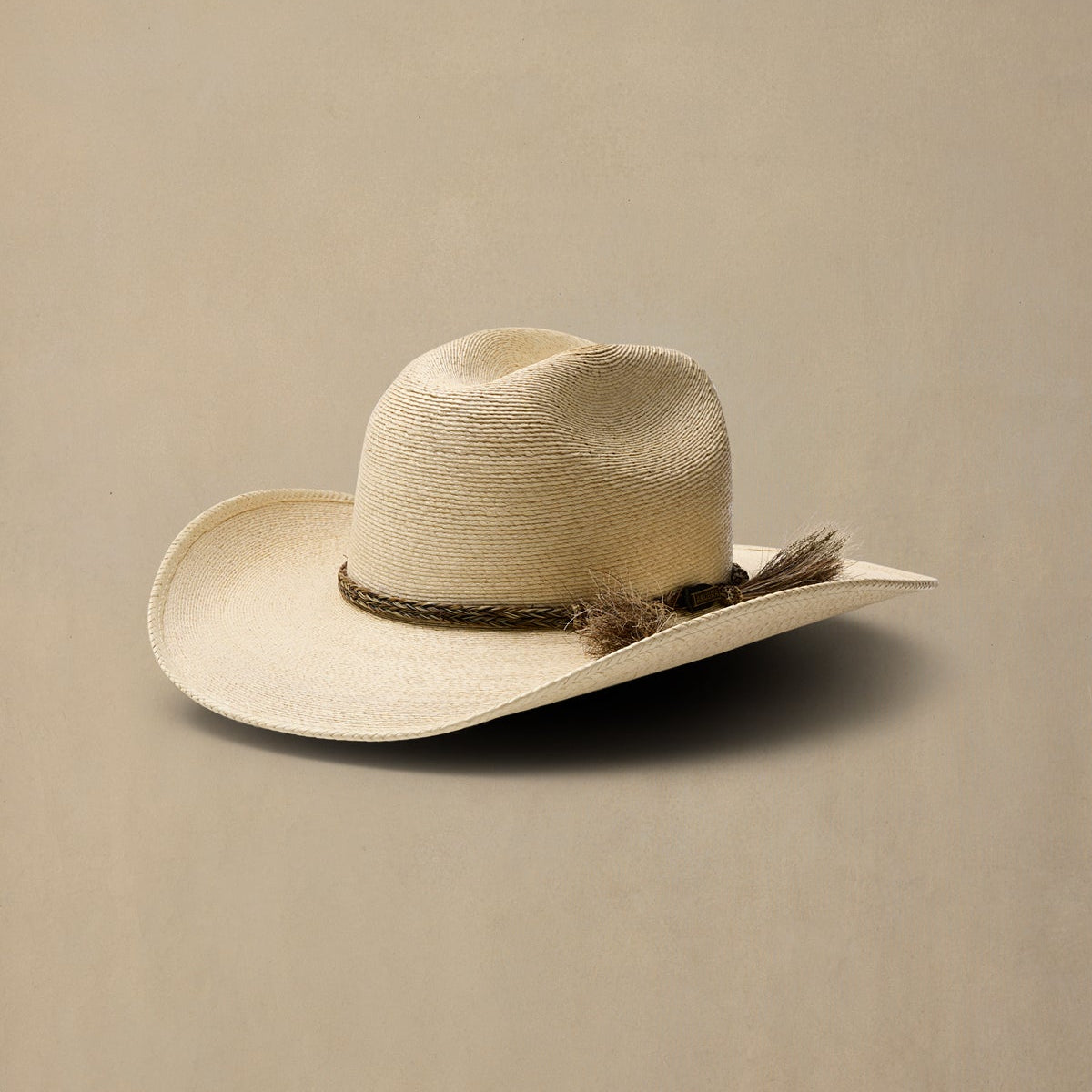 A light beige straw cowboy hat with a dark brown woven band and tassel accent, resting on a solid beige background.