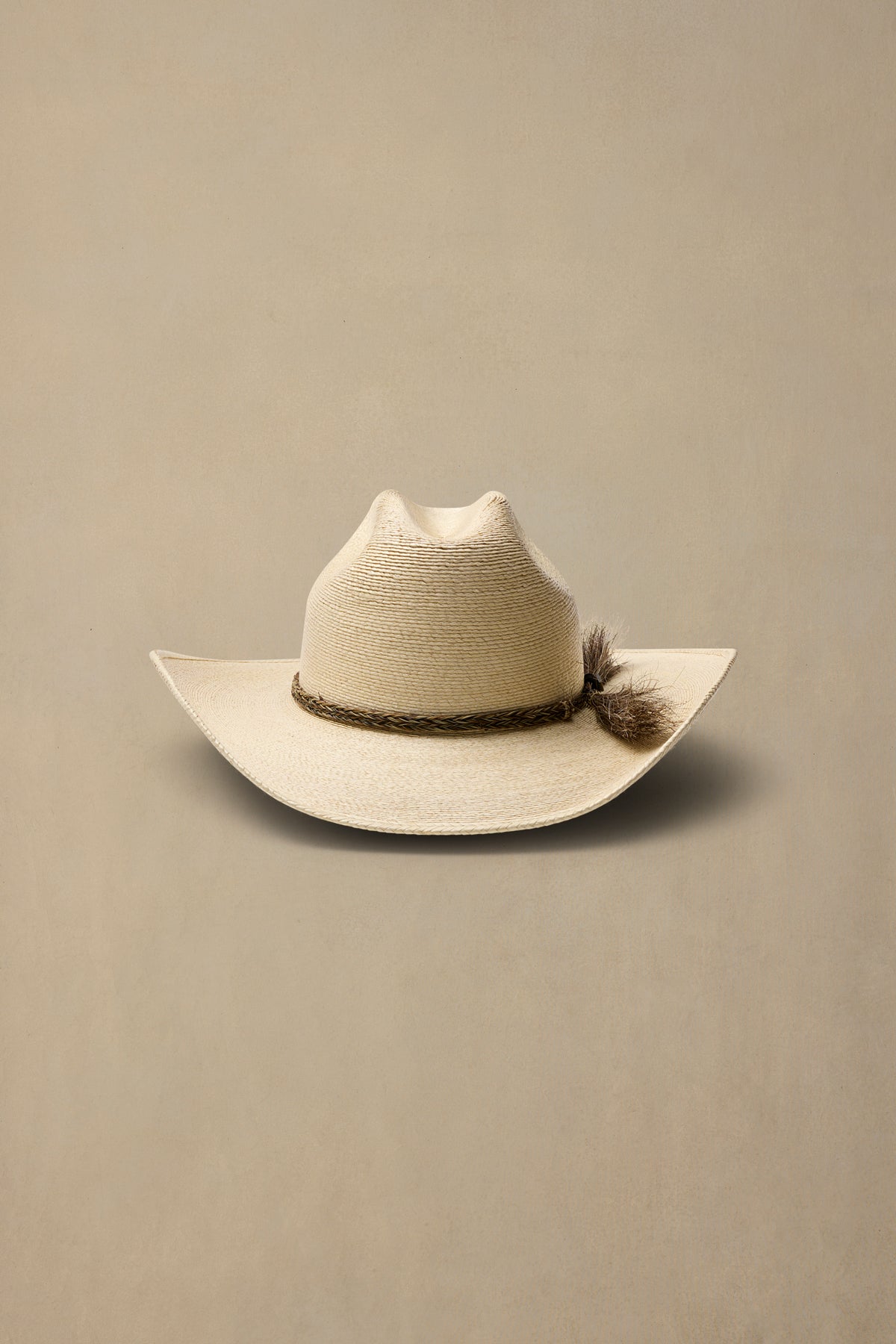 A light beige straw cowboy hat with a dark brown braided band and tassel accent, photographed against a solid beige background.