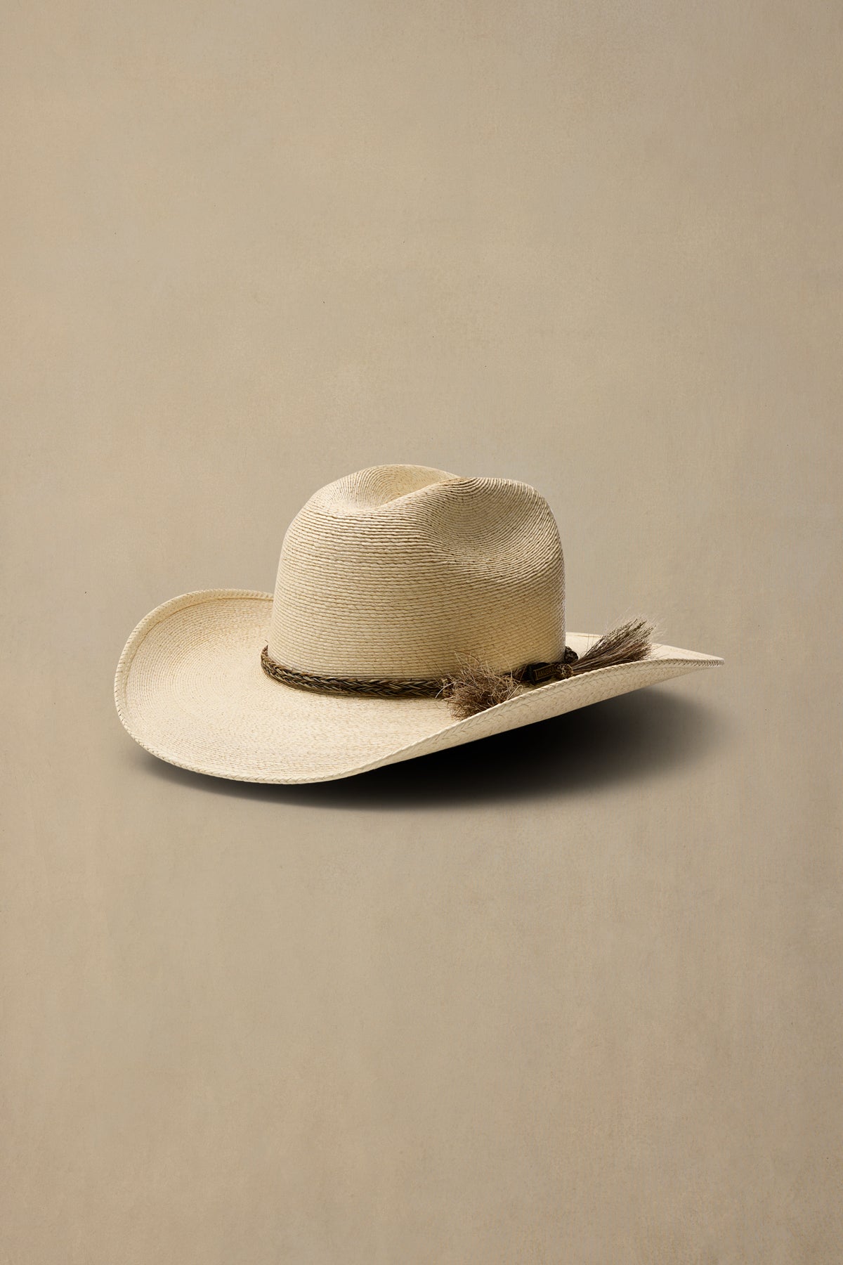 A light beige straw cowboy hat with a dark brown woven band and tassel accent, resting on a solid beige background.