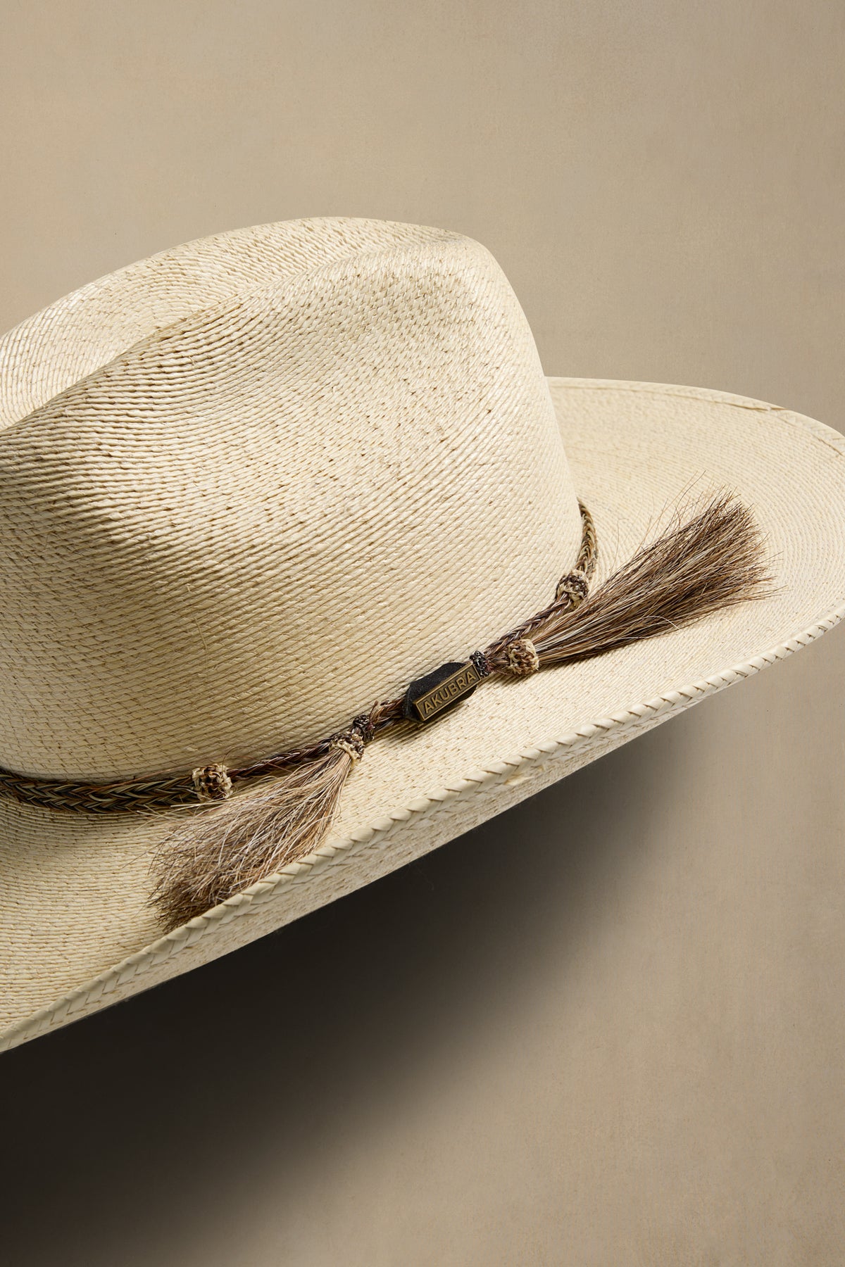 A close-up of a woven straw cowboy hat in a light beige color, featuring a brown braided band with two brown tassels, set against a solid neutral beige background.