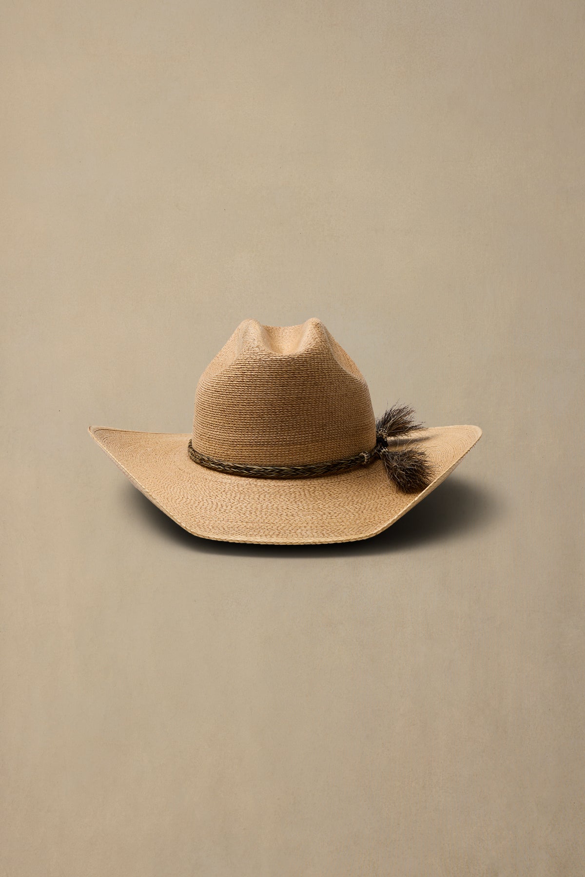 A dark tan woven cowboy hat with a dark brown braided band and a tassel accent on the side, displayed against a solid beige background.