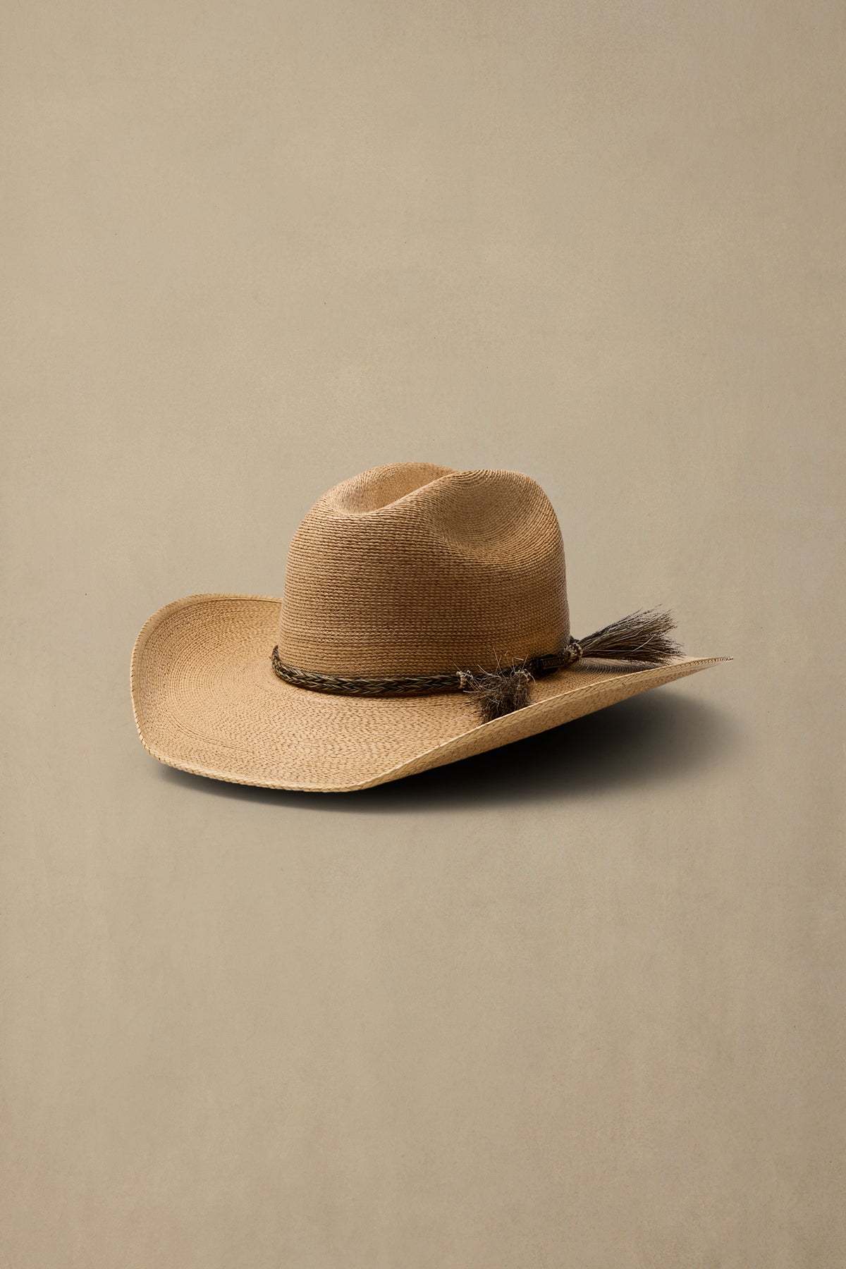 A dark tan woven cowboy hat with a dark brown decorative band and tassel accent, set against a solid beige background.