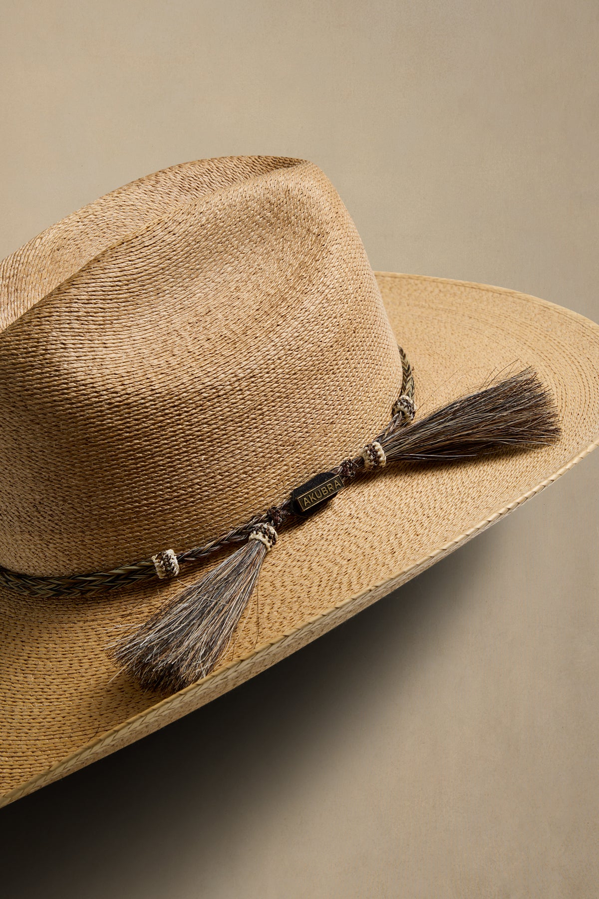A close-up of a woven straw hat in a dark tan colour, featuring a dark brown braided band with matching brown tassel, set against a solid beige background.