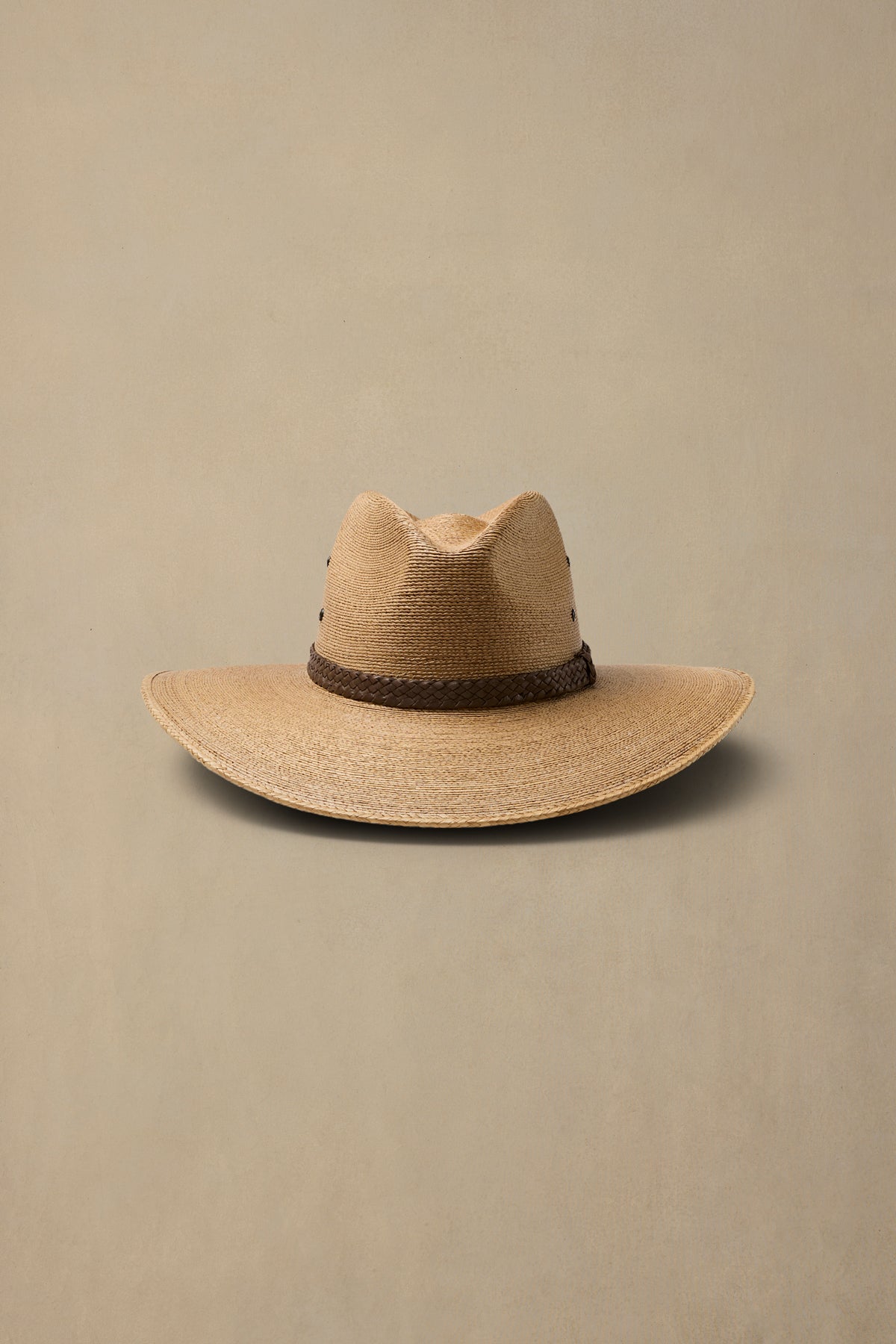 A dark tan woven straw cowboy hat with a dark brown braided band, displayed against a solid beige background.
