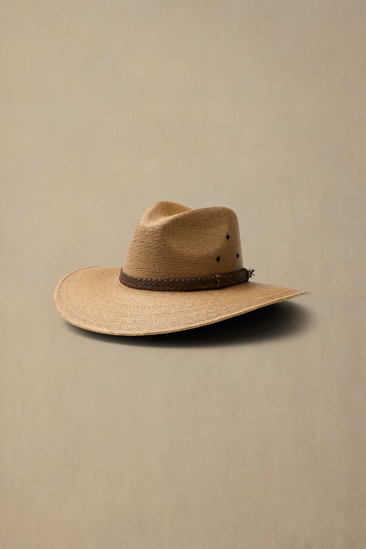 A dark tan woven cowboy hat with a dark brown braided band, photographed on a solid beige background.