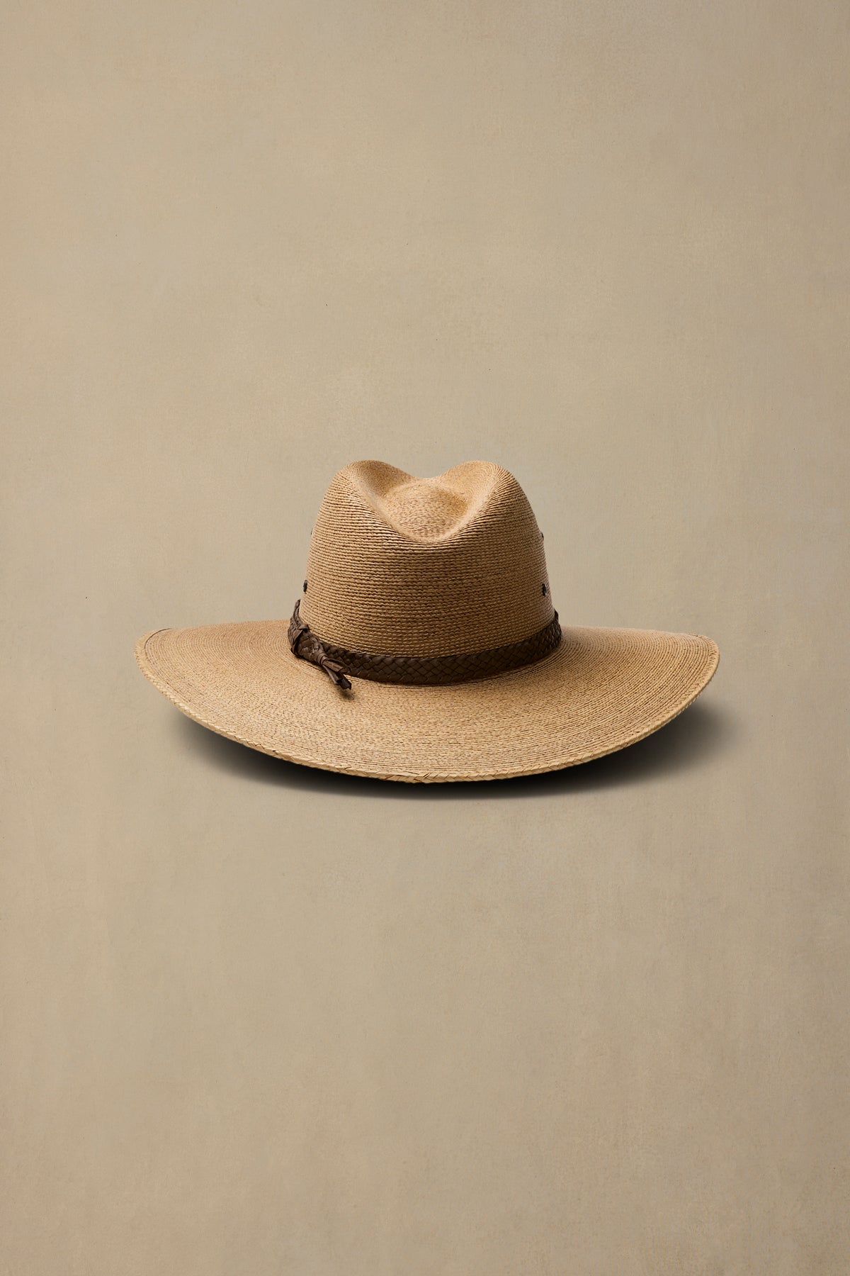A dark tan woven hat with a dark brown band, photographed against a solid beige background.