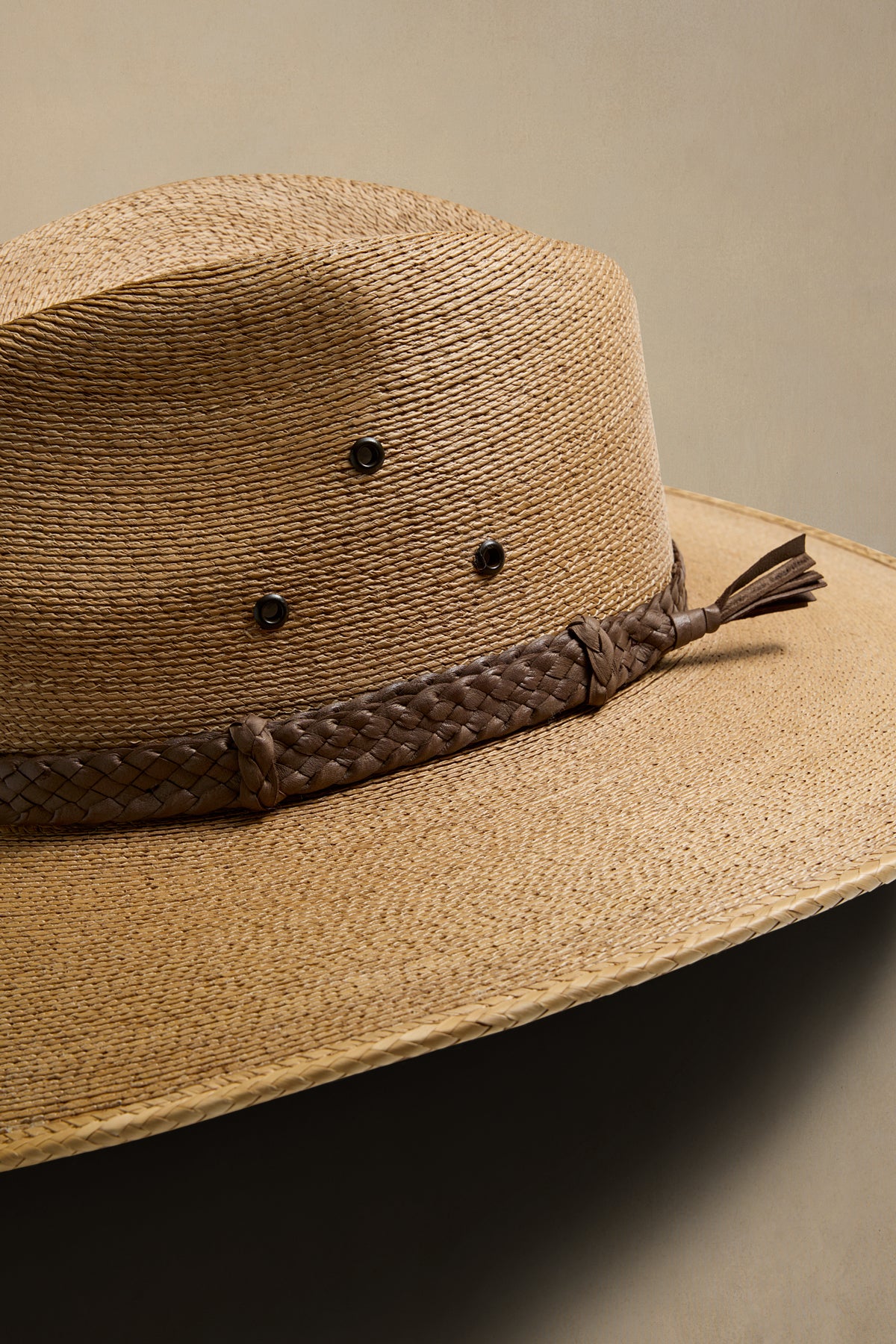 A close-up of a woven straw country hat in a dark tan color, featuring a dark brown braided band with a tassel and three small decorative studs on the crown, set against a solid beige background.