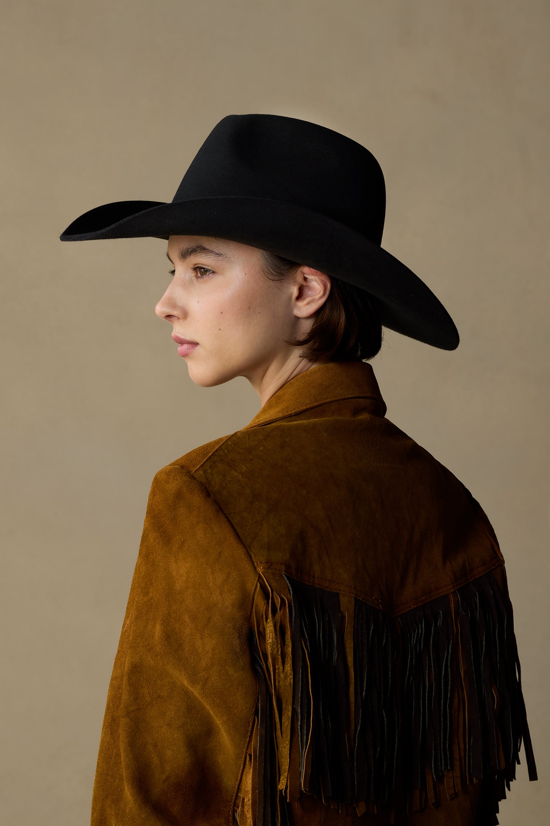 Black cowboy hat with brown suede fringe jacket, side profile view against neutral tan background