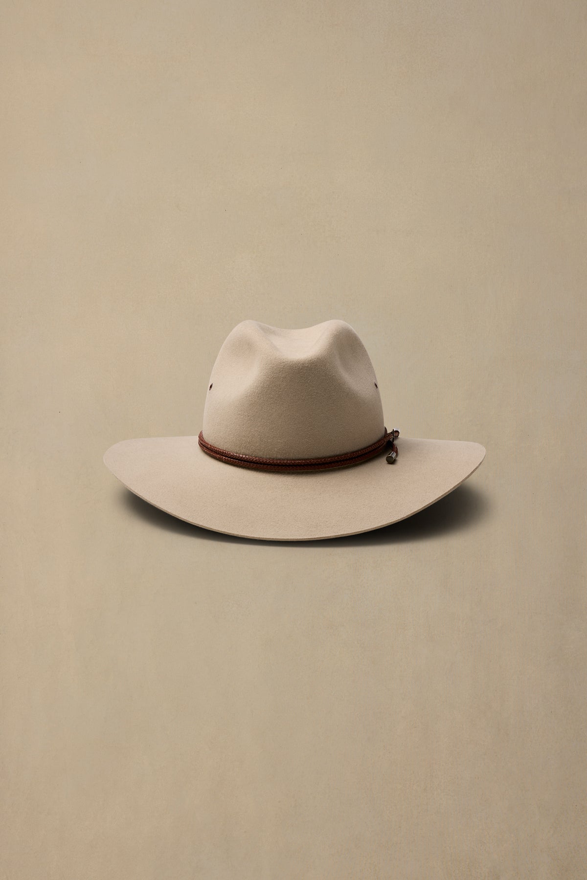 Cream-colored rabbit fur felt cowboy hat with brown cord band on neutral beige background