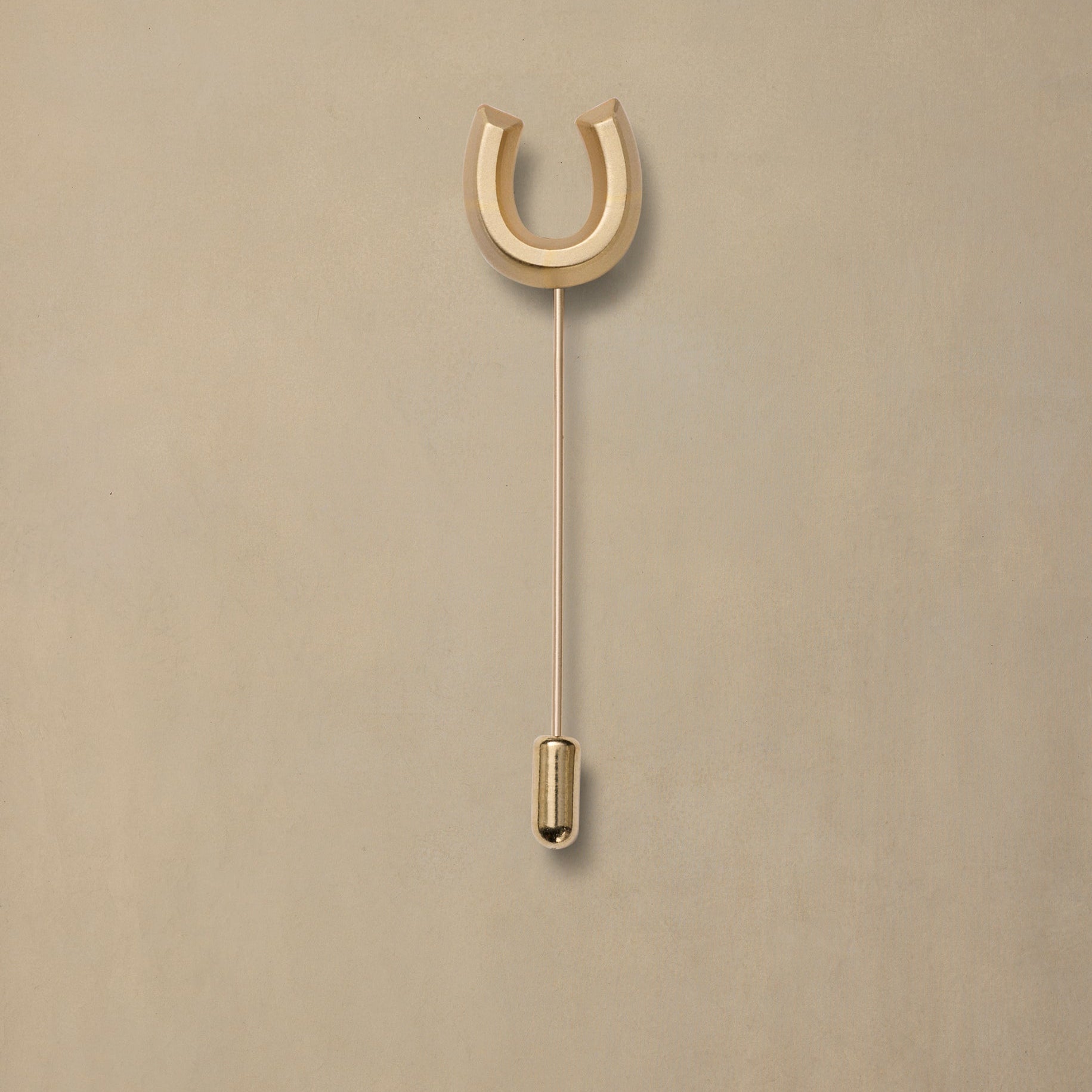 Polished brass horseshoe pin with thin stem and rounded gold cap on neutral beige background