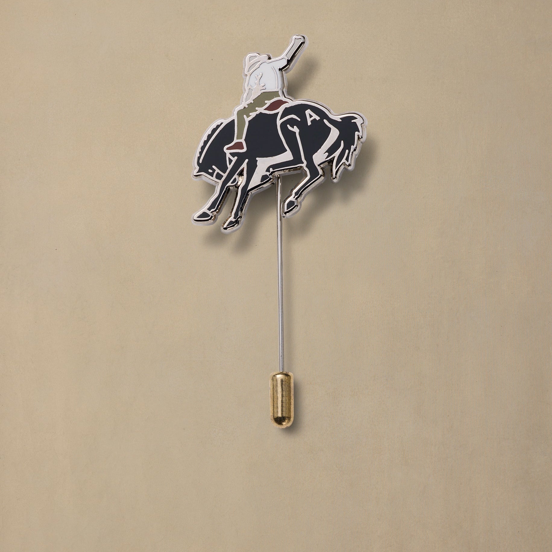 Bucking bronc enamel pin with white horse rider on black horse, gold clutch backing, displayed against beige background