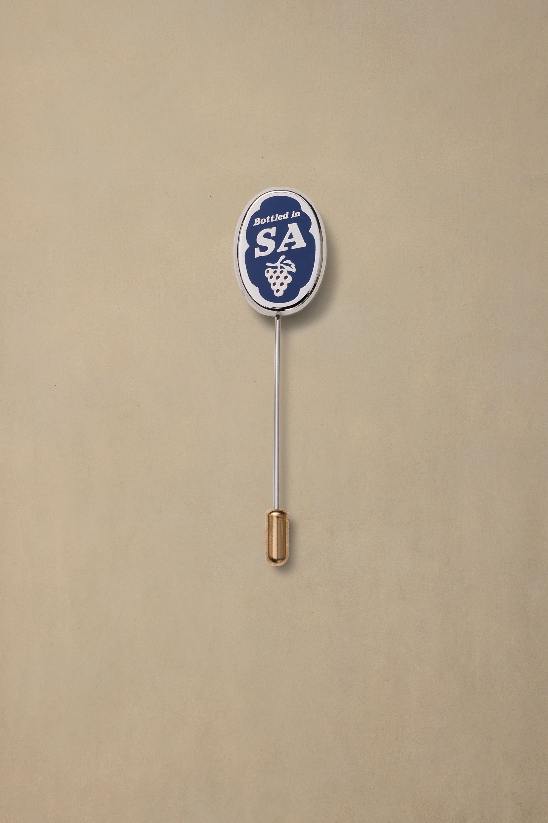 Navy and white enamel pin with "Bottled in SA" text and grape cluster design, gold-tone pin back on beige background