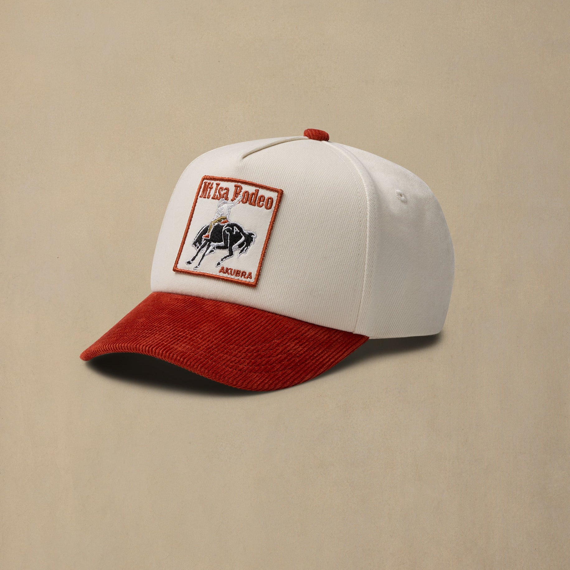 A white baseball cap with a red corduroy brim and a rectangular 'Mt Isa Rodeo 2026' patch on the front, set against a solid beige background.