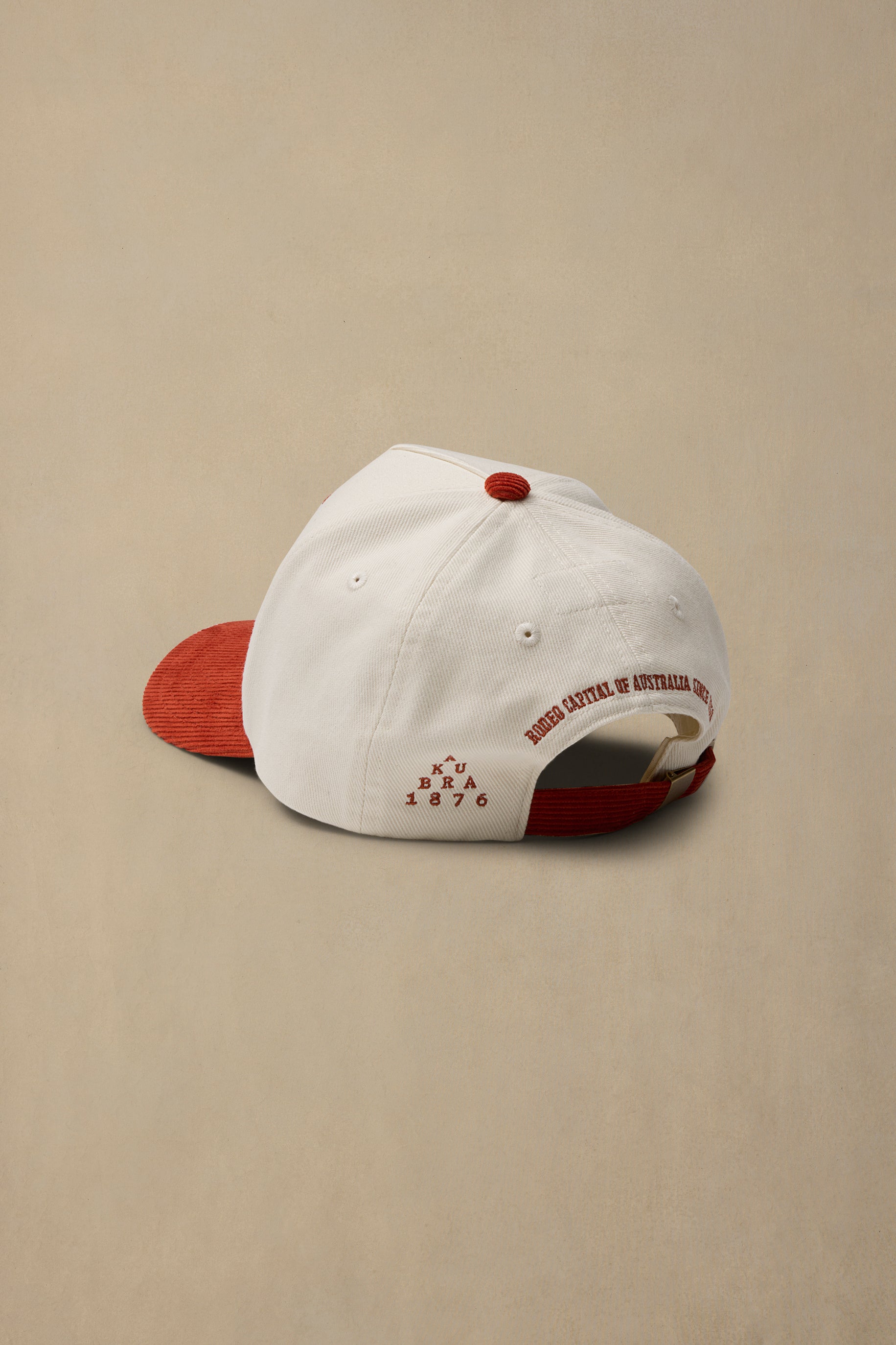 A white baseball cap with a red visor and red back strap, shown from a side-back angle on a warm beige background. The white crown features red embroidered text reading 'RODEO CAPITAL OF AUSTRALIA MT ISA' and '1876' with a small red geometric logo.