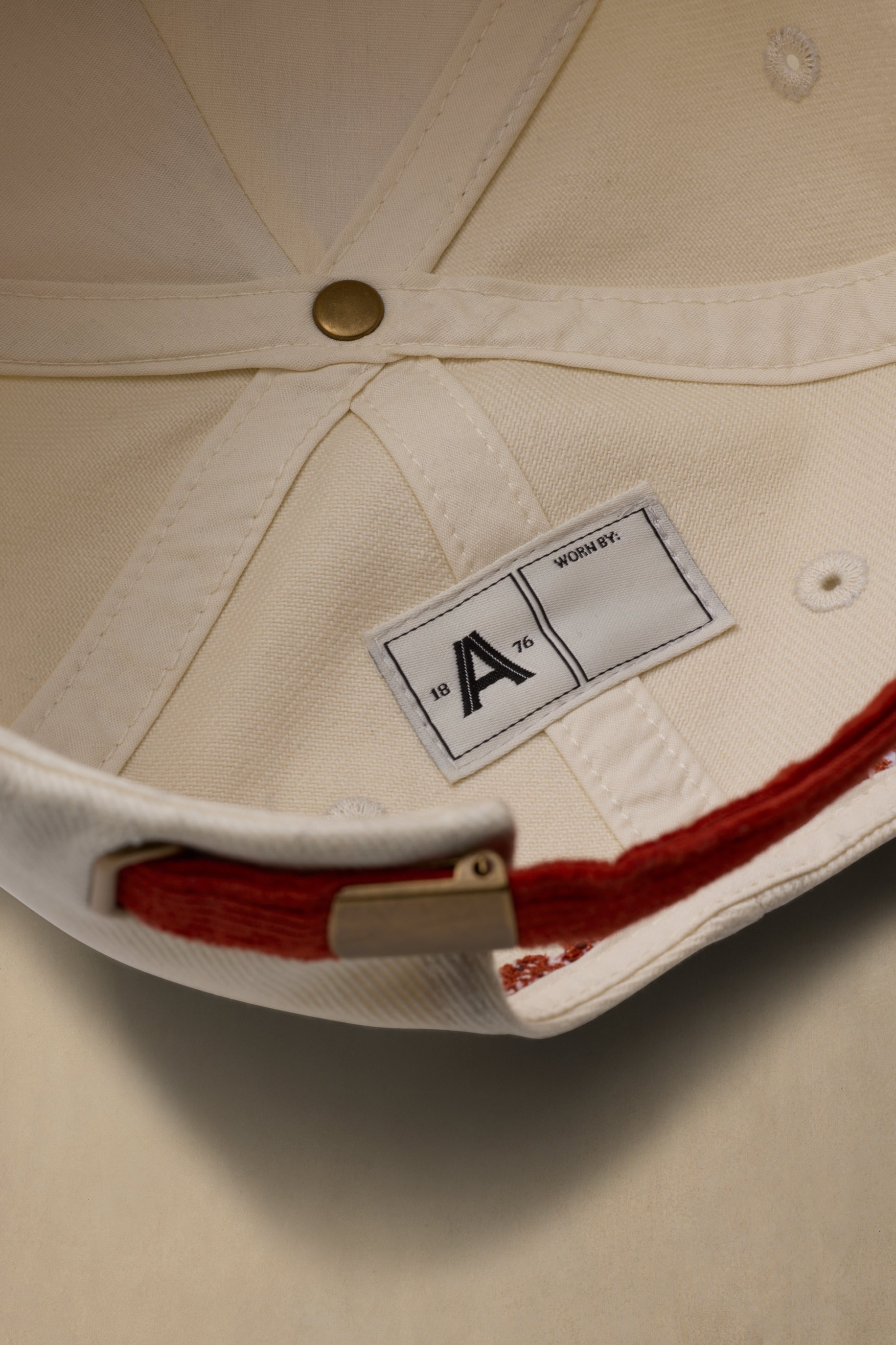 Close-up of the inside crown of a cream-colored baseball cap, showing the fabric construction, a white size label with a black letter 'A', and a red stitched detail along the brim edge.
