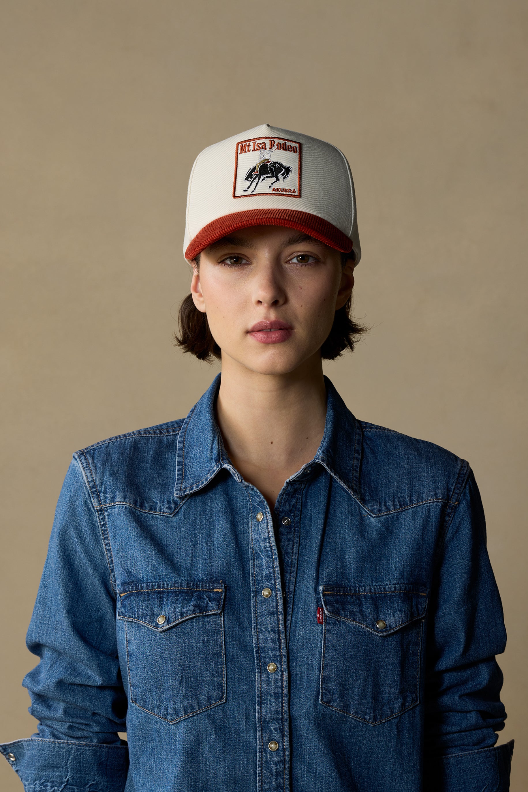 A model wearing a white and red trucker hat with a 'Mt Isa Rodeo' patch and a blue denim shirt against a beige background.