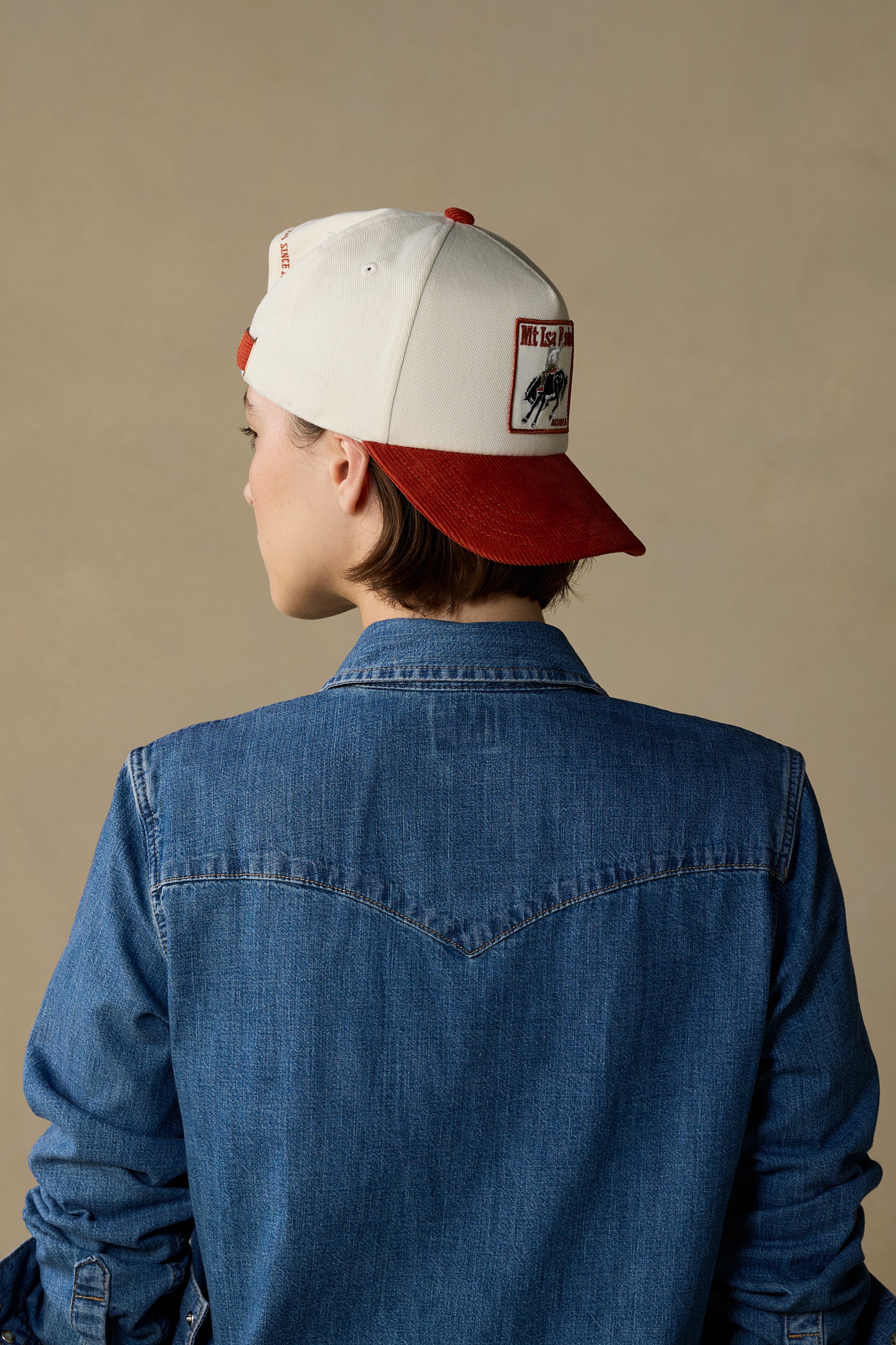 A person shown from the back wearing a white and red baseball cap with a patch on the side, along with a blue denim shirt, against a plain beige background.