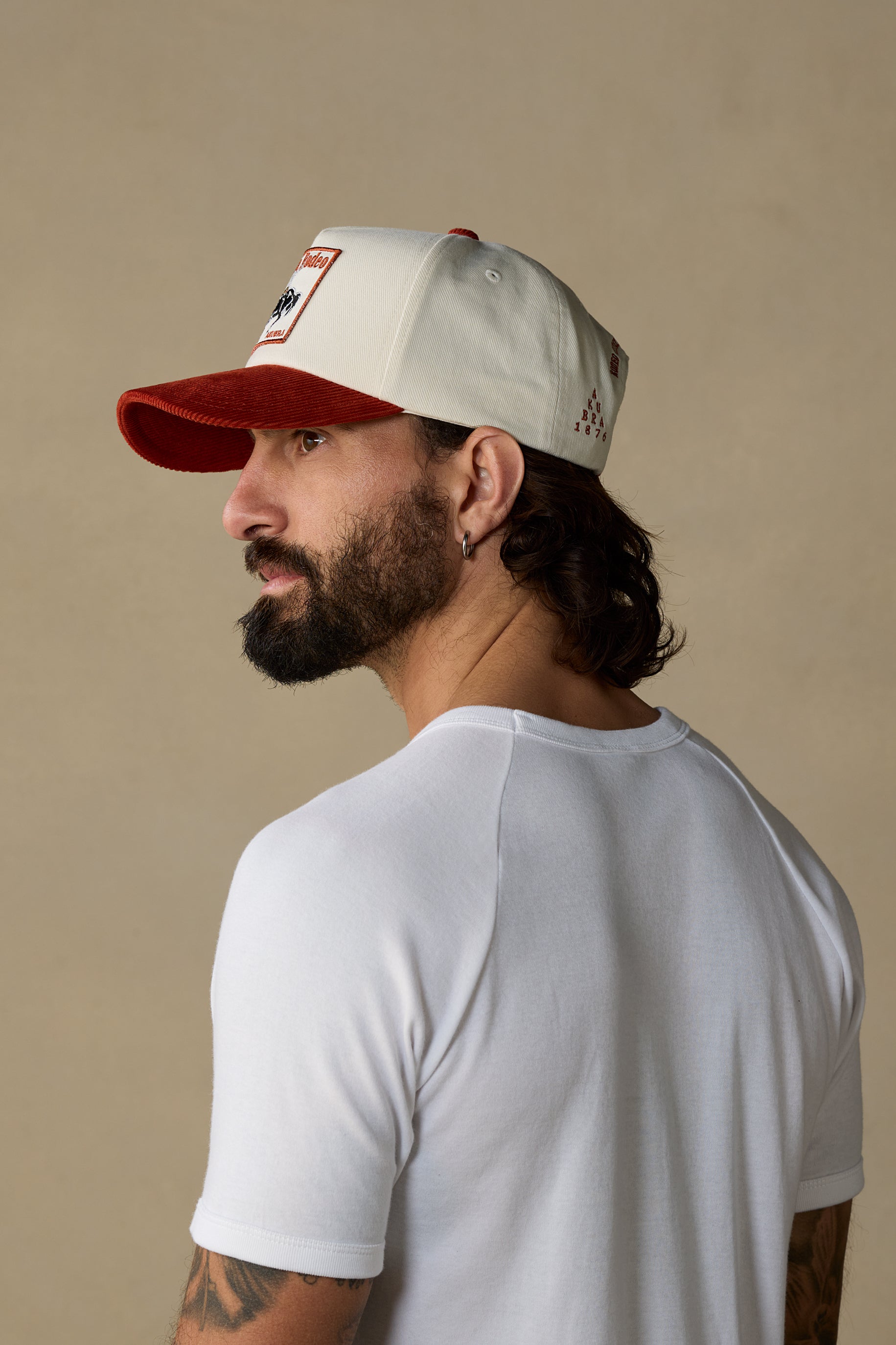 A man shown from the back and side profile wearing a white baseball cap with a red brim and a small red-and-black logo patch on the front, paired with a white short-sleeve shirt against a plain beige studio background.