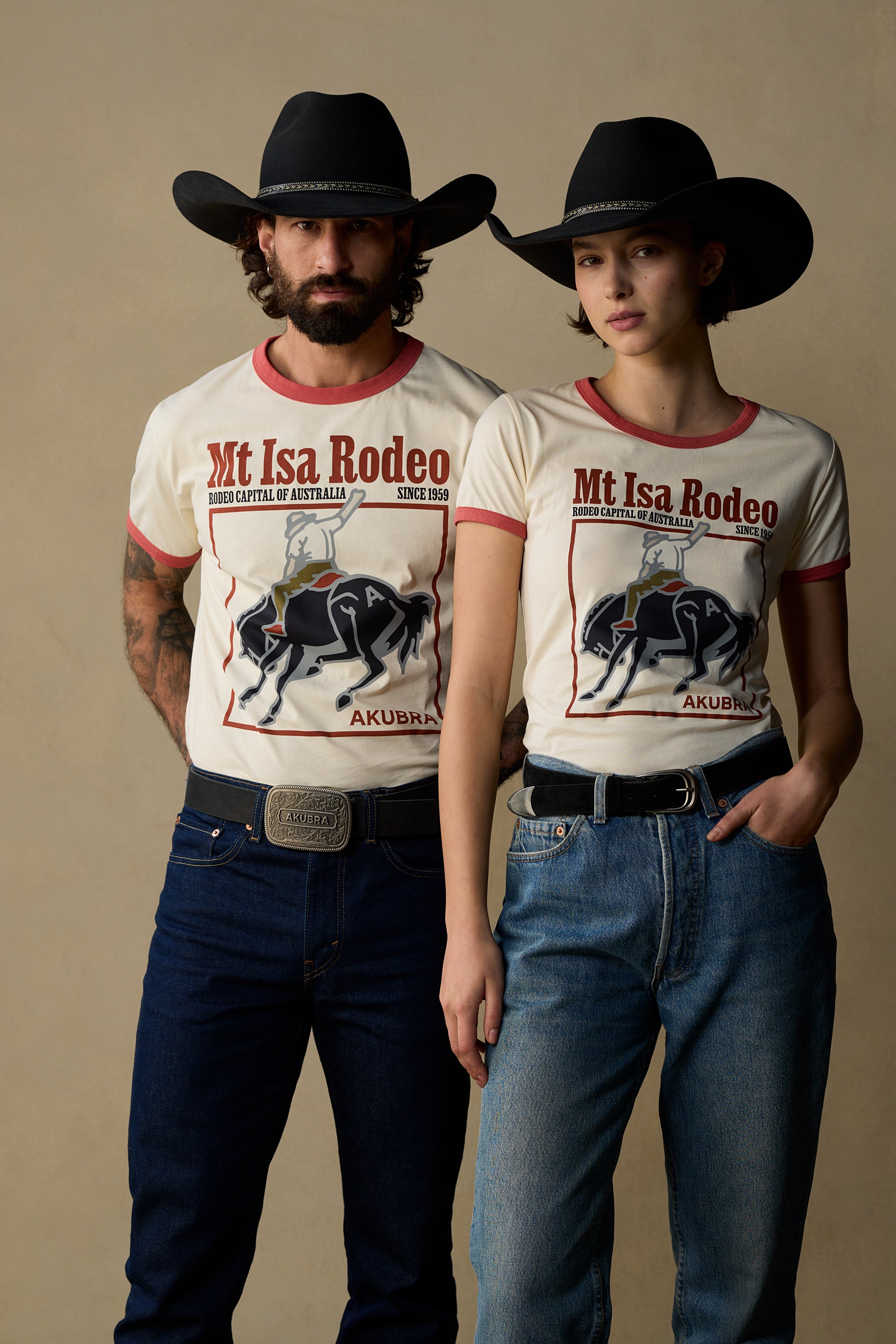 A man and a woman wearing matching cream ringer T-shirts with a Mt Isa Rodeo graphic, paired with black cowboy hats, dark blue jeans, and black belts, standing against a solid beige studio background.