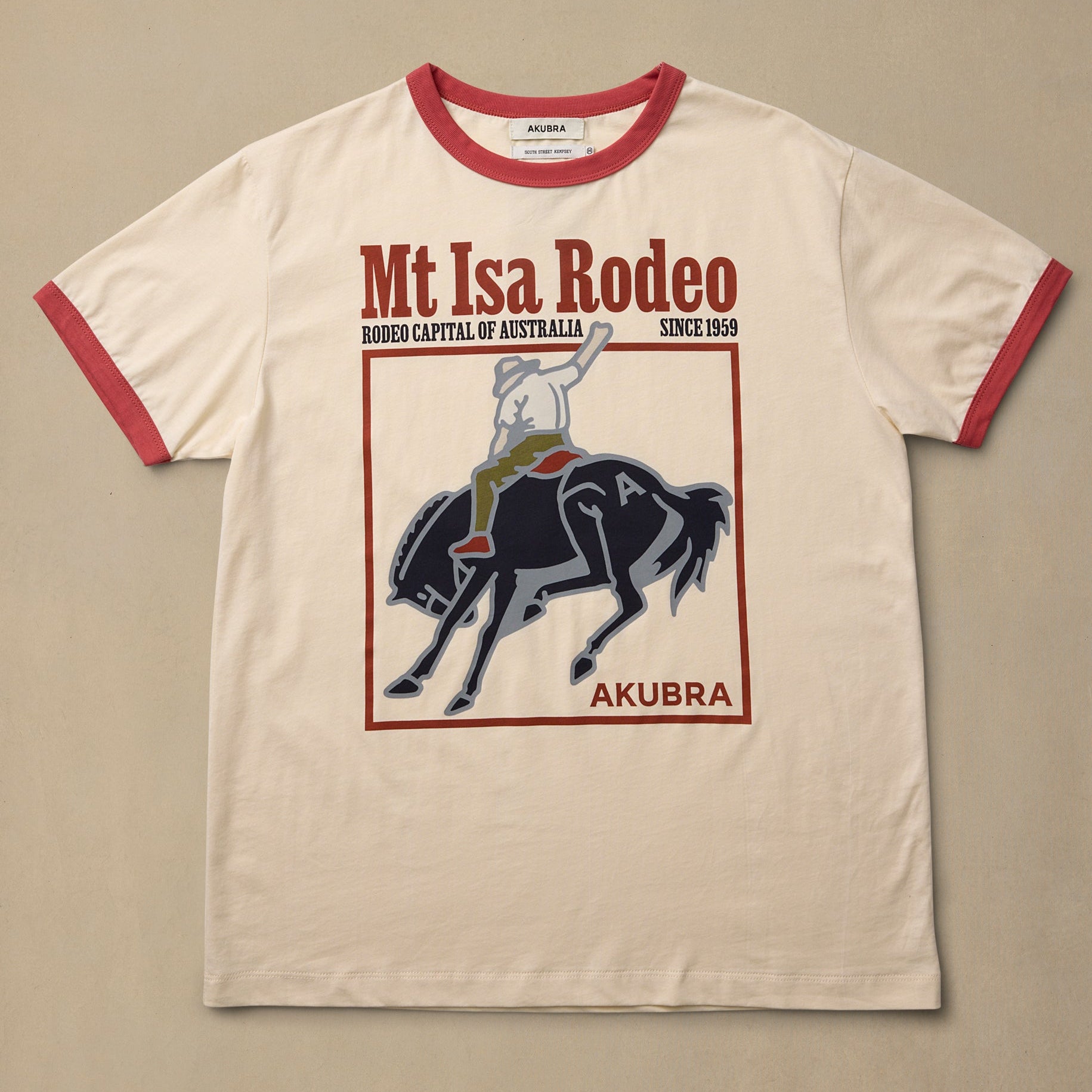 A cream-colored ringer t-shirt with red trim on the collar and sleeves, featuring a graphic print of a cowboy riding a bucking horse with the text 'Mt Isa Rodeo' and 'Akubra'.