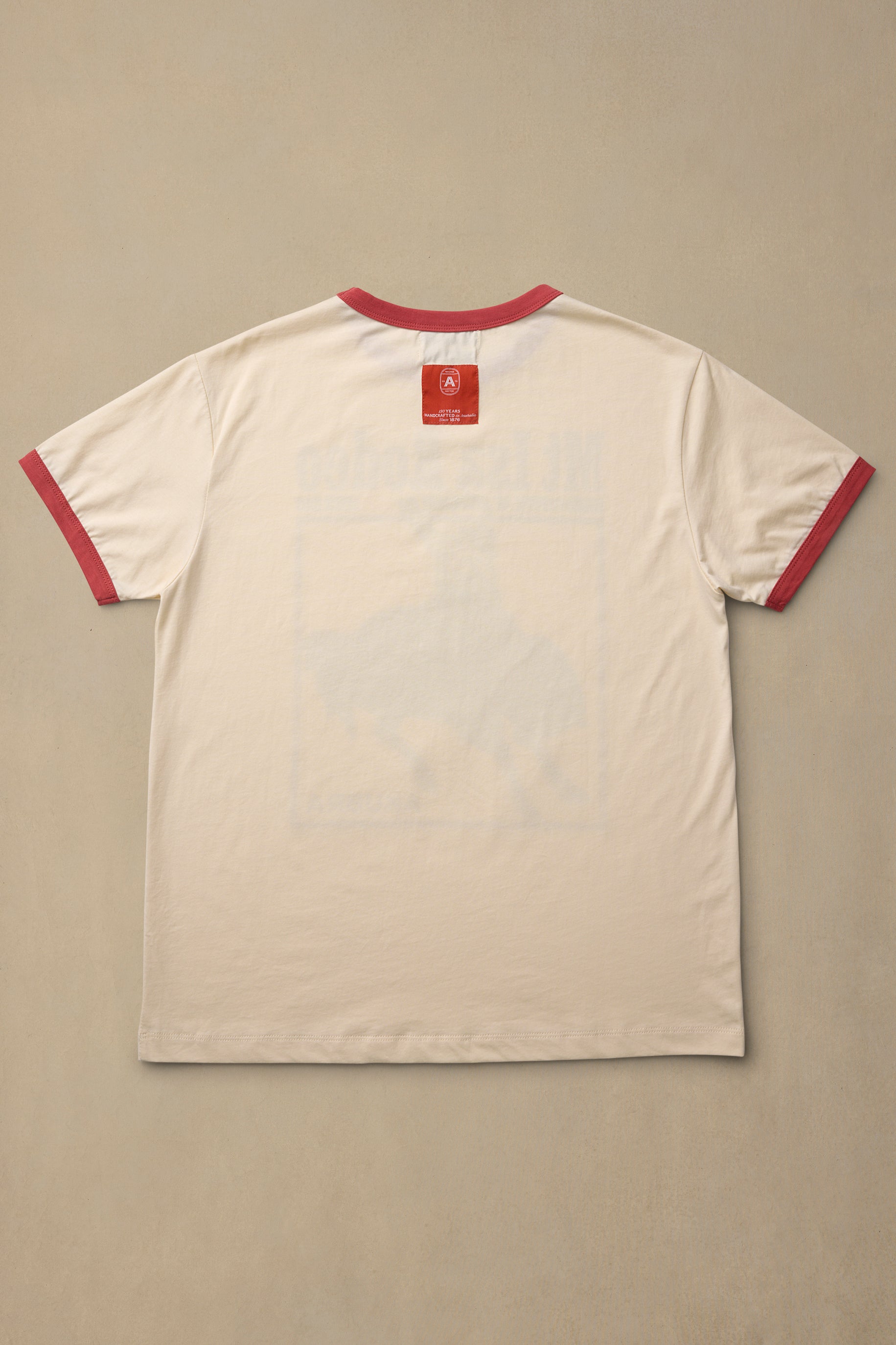 A cream-colored ringer t-shirt with red trim on the collar and sleeve cuffs, laid flat against a neutral beige background.