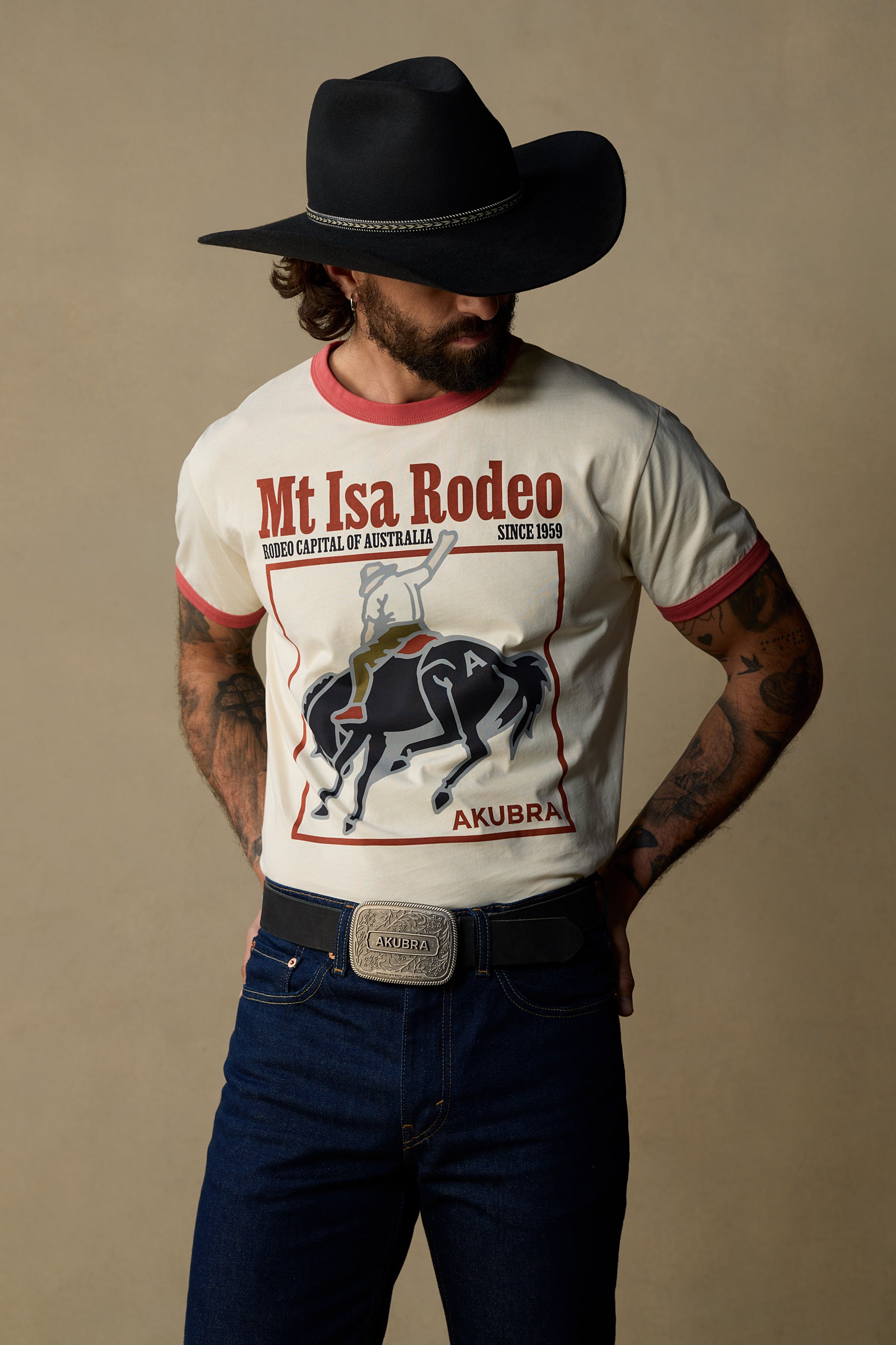A man wearing a black cowboy hat, a cream ringer t-shirt with a red and black Mt Isa Rodeo graphic, and dark blue jeans, posed against a solid beige studio background.