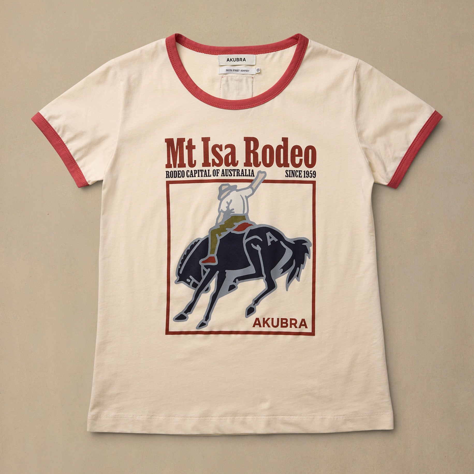 An off-white ringer t-shirt with red trim on the collar and sleeves, featuring a graphic print of a rider on a horse and the text 'Mt Isa Rodeo' and 'AKUBRA'.