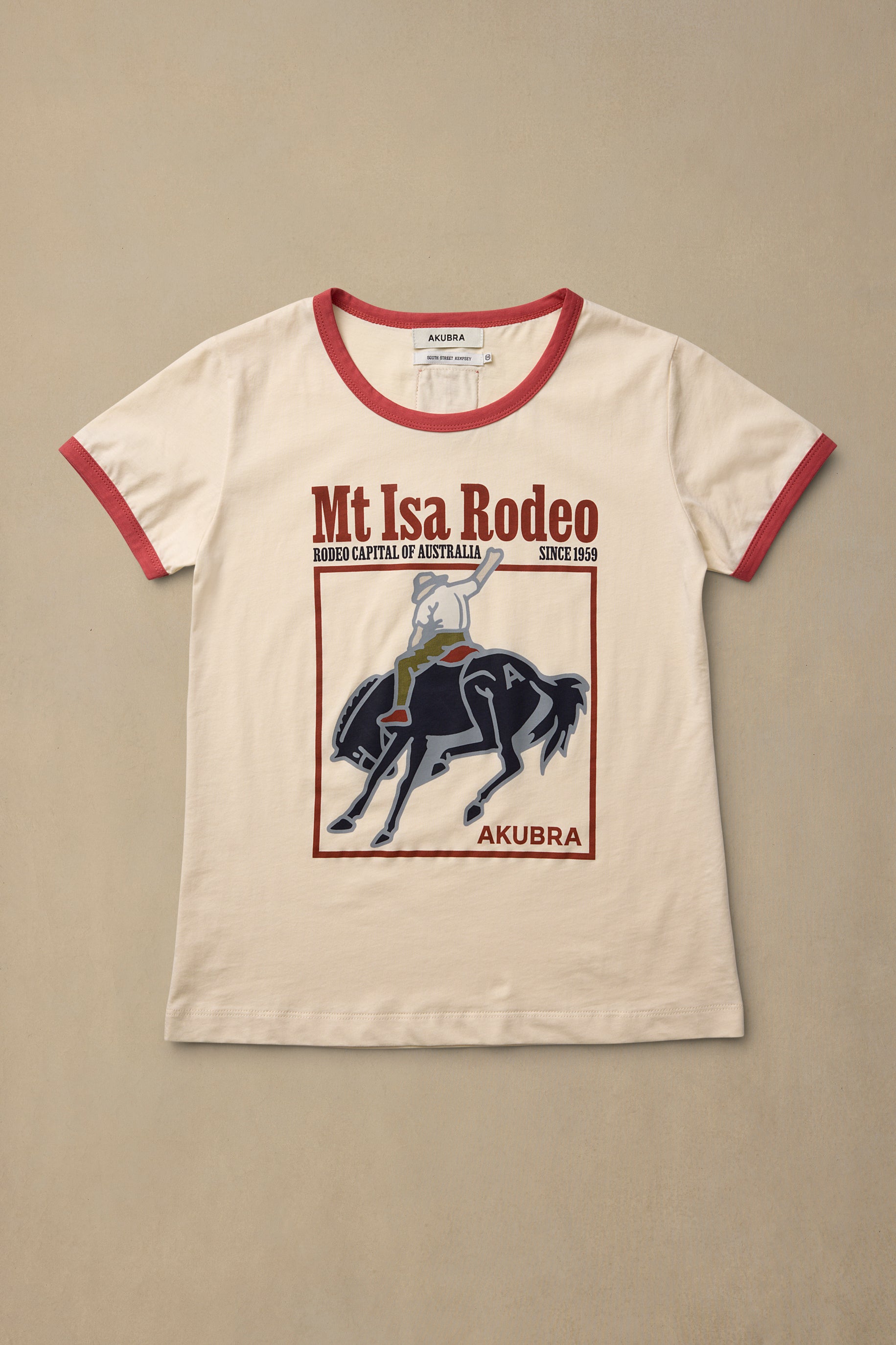 An off-white ringer t-shirt with red trim on the collar and sleeves, featuring a graphic print of a horse rider and the text 'Mt Isa Rodeo' and 'AKUBRA'.