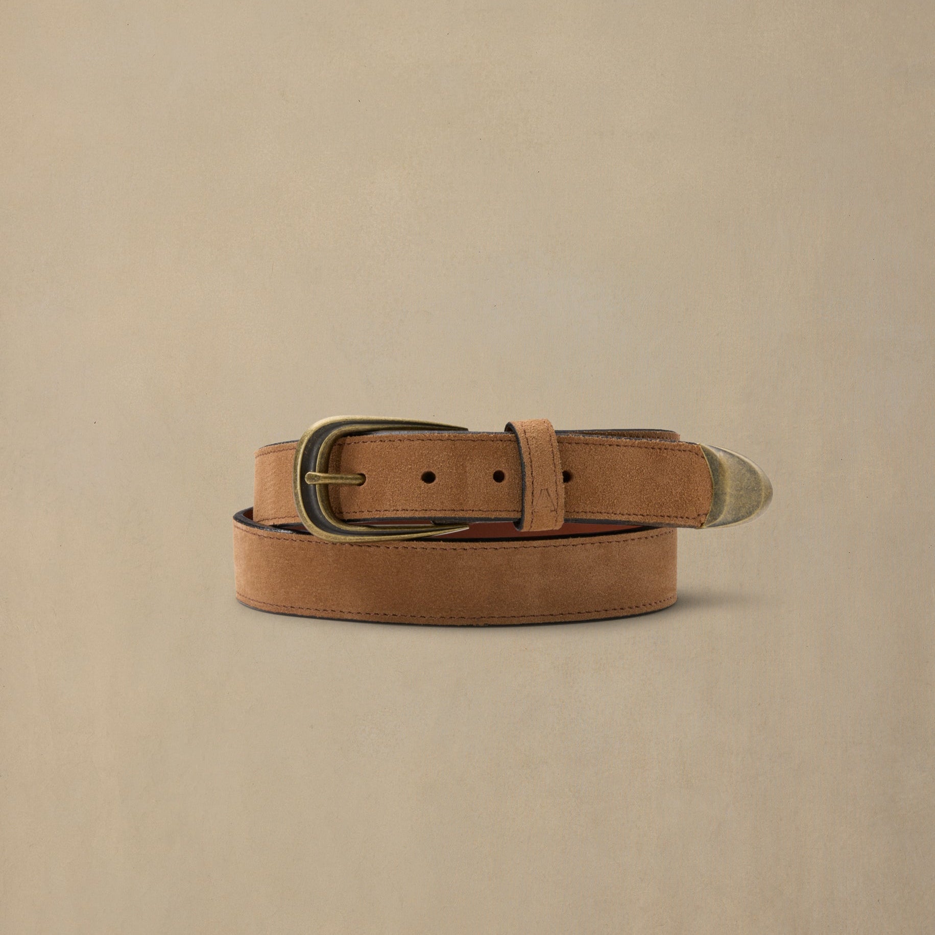Tan suede belt with brass buckle and metal tip detail on neutral background
