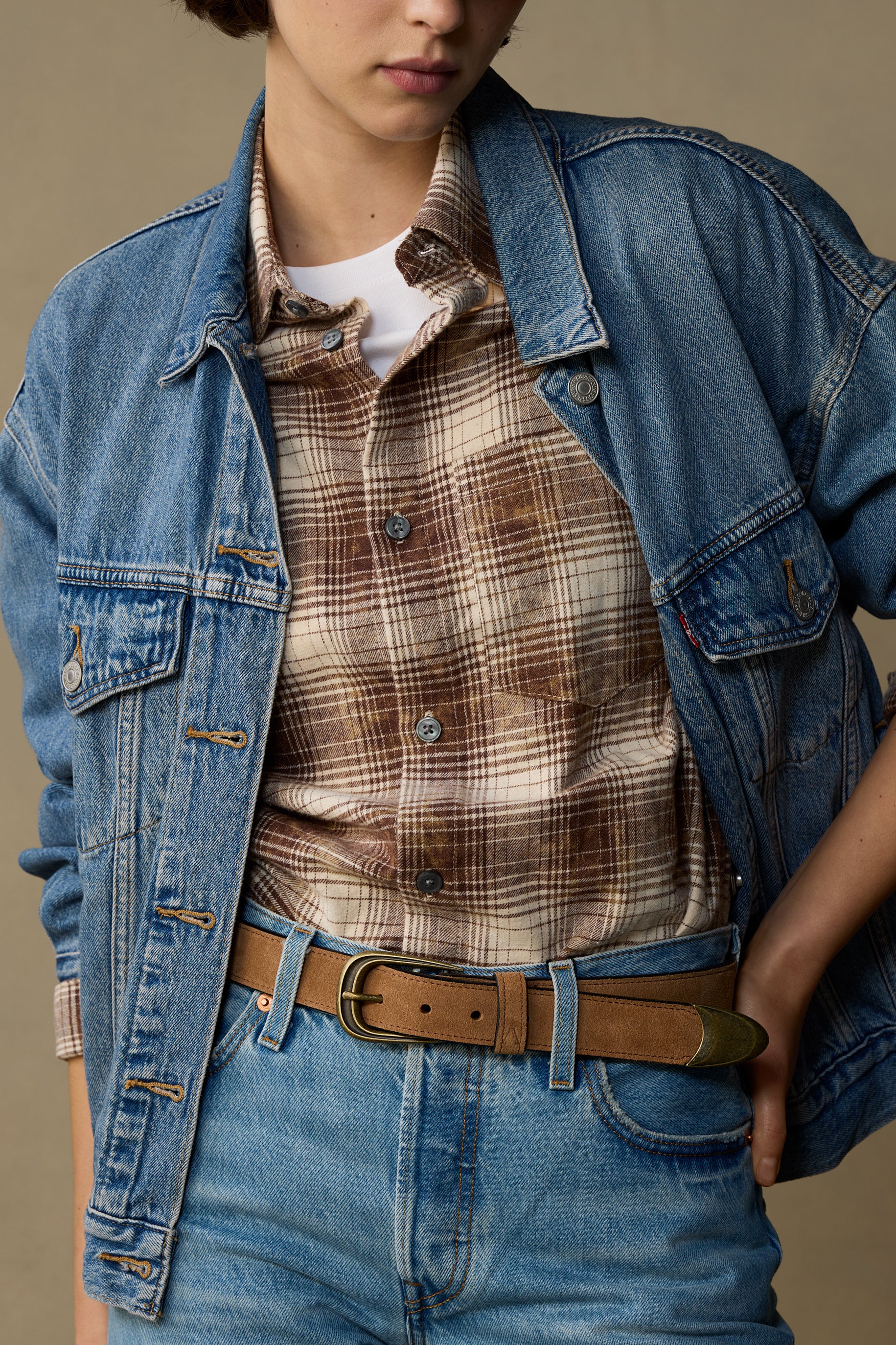 Tan suede belt with brass buckle worn with denim jacket and plaid shirt on neutral background