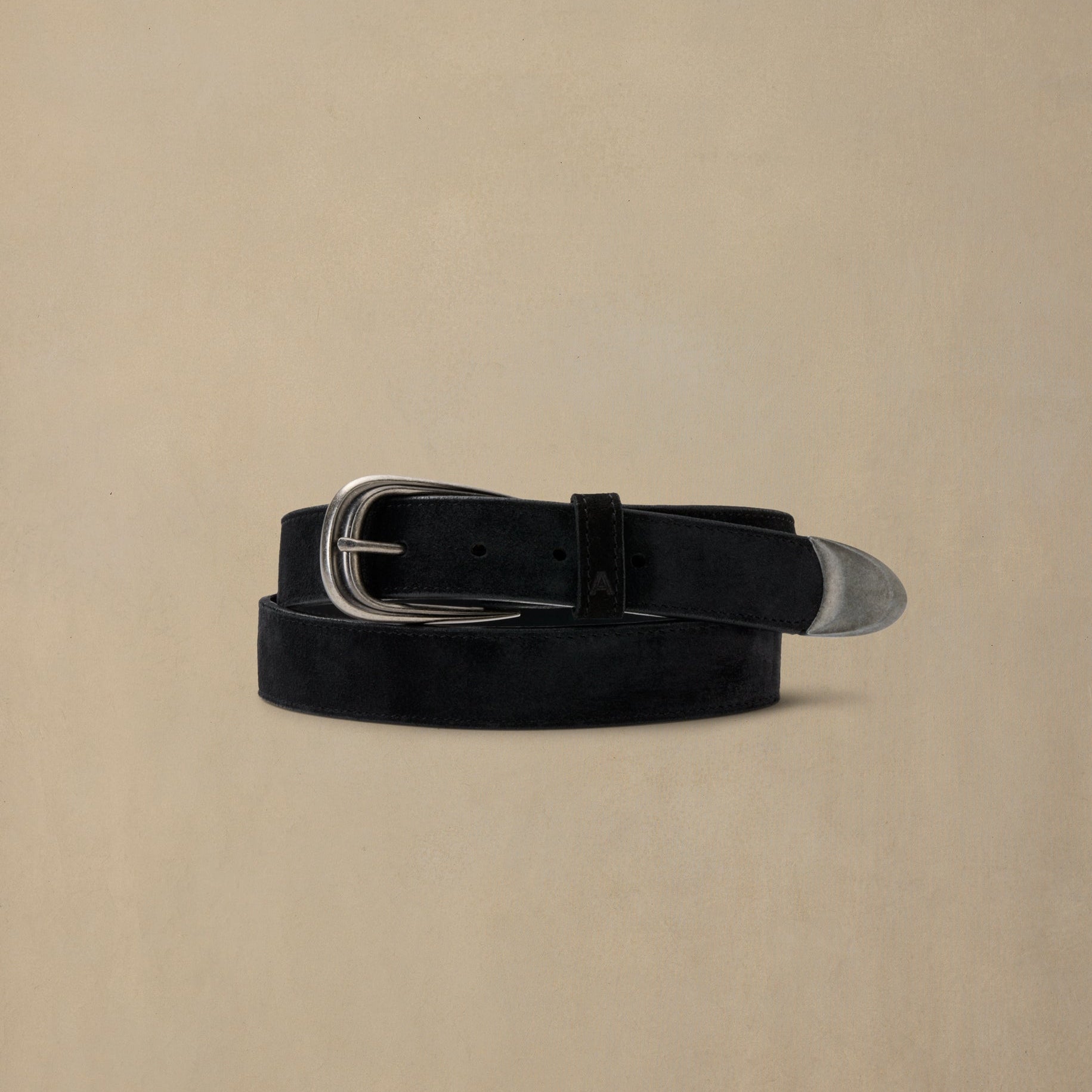 Black suede belt with silver oval buckle and metal tip, coiled on neutral beige background