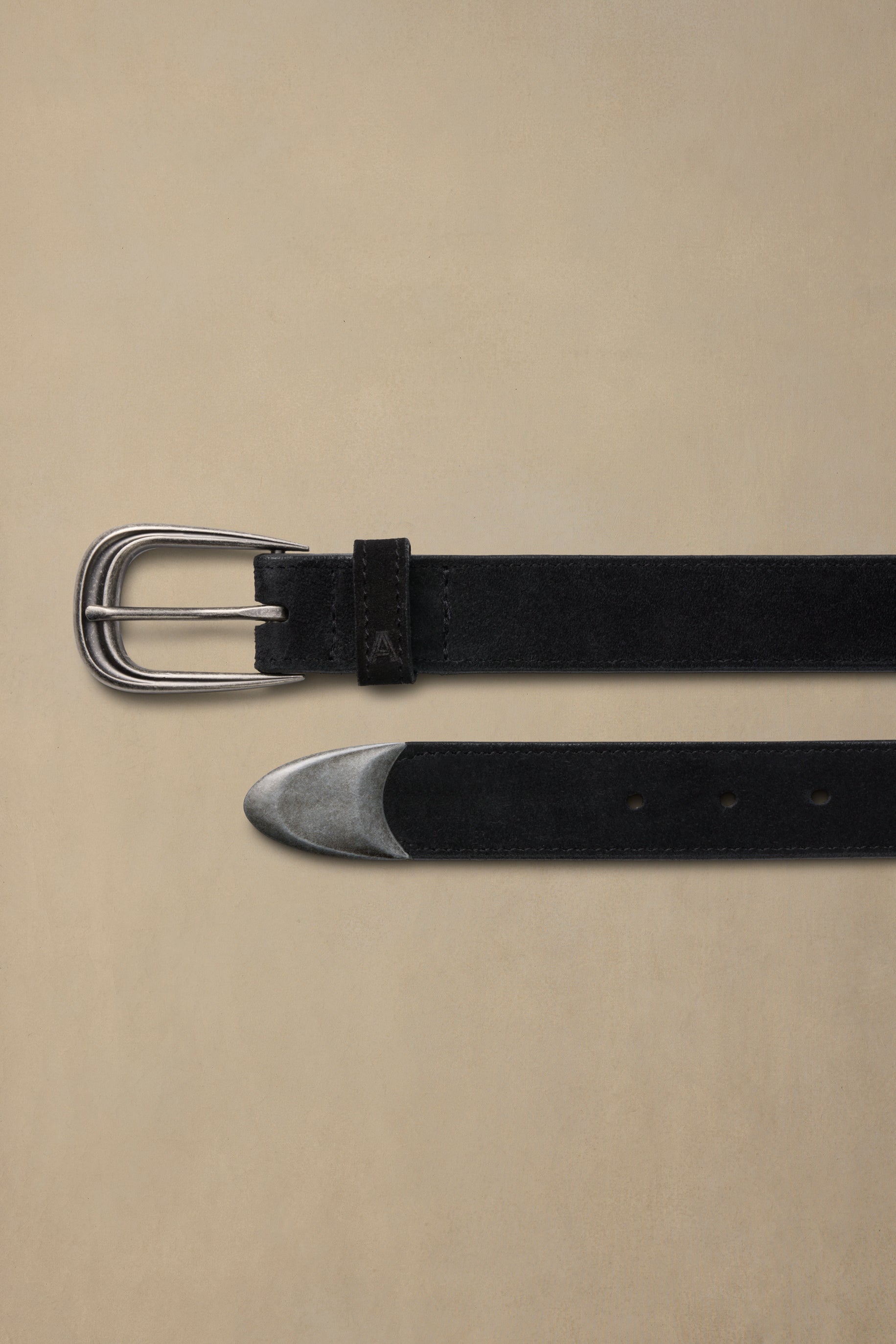 Black suede belt with pewter buckle and metal tip displayed flat on beige background