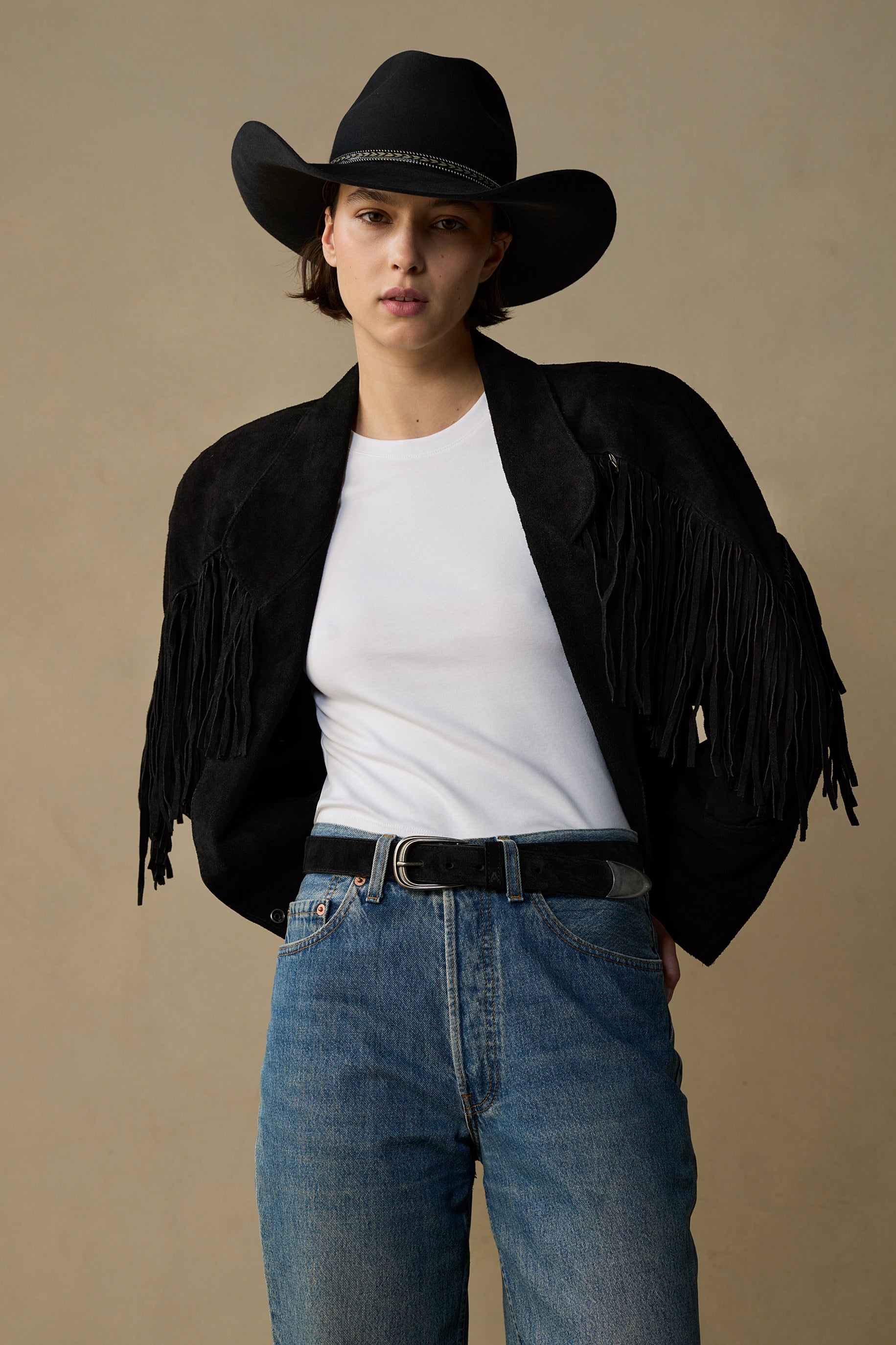 Black suede belt with pewter buckle worn with white tee, fringe jacket and cowboy hat against neutral background
