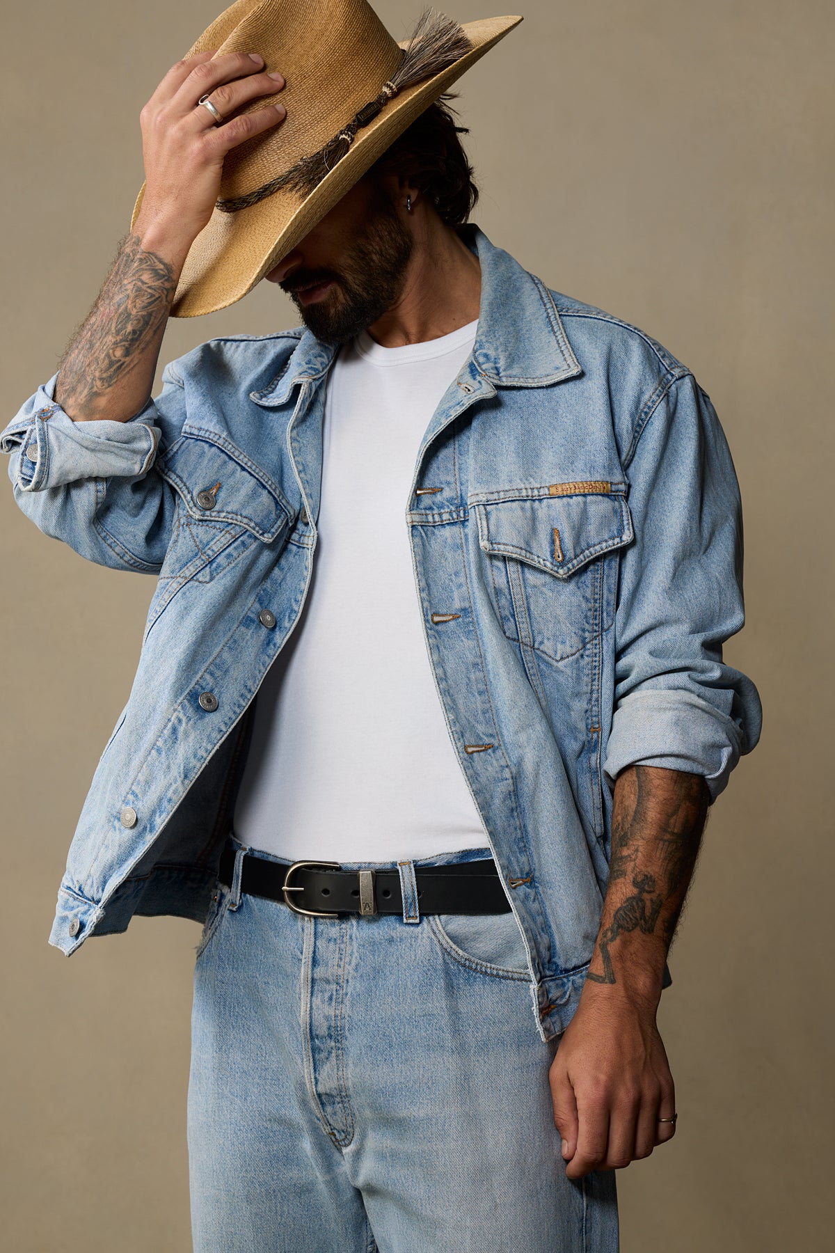 Man wearing light wash denim jacket and jeans with black leather belt against neutral tan background