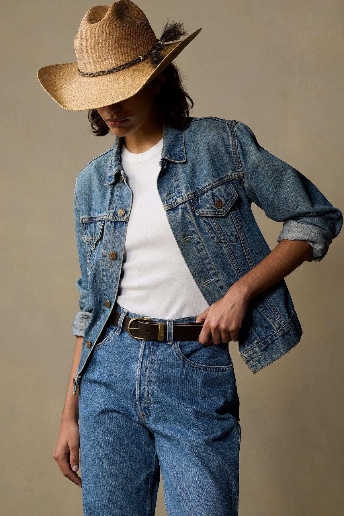 Chocolate brown leather belt with brass buckle worn with denim jacket and cowboy hat on neutral background