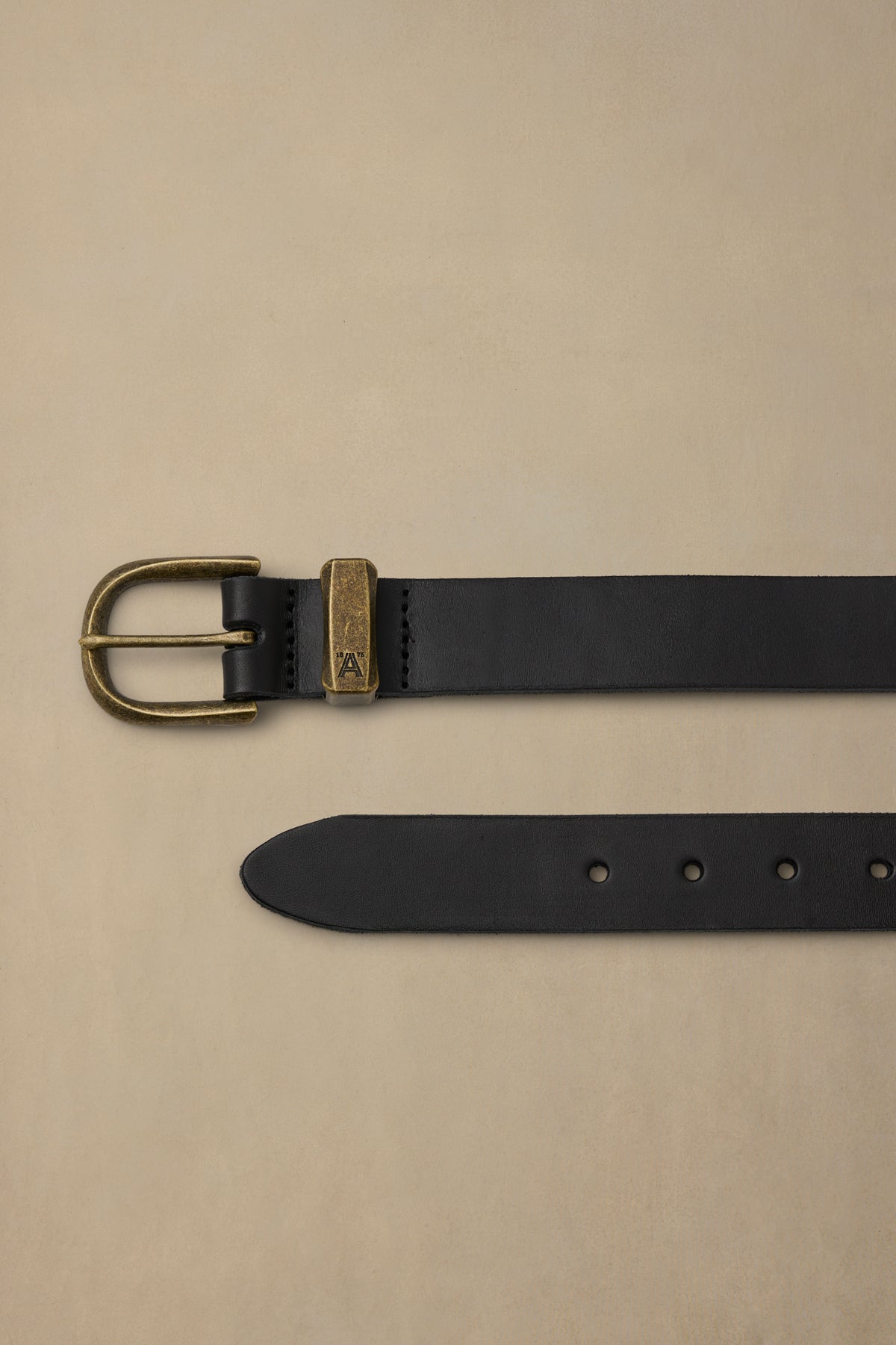Black leather belt with brass buckle and keeper loop displayed flat on beige background