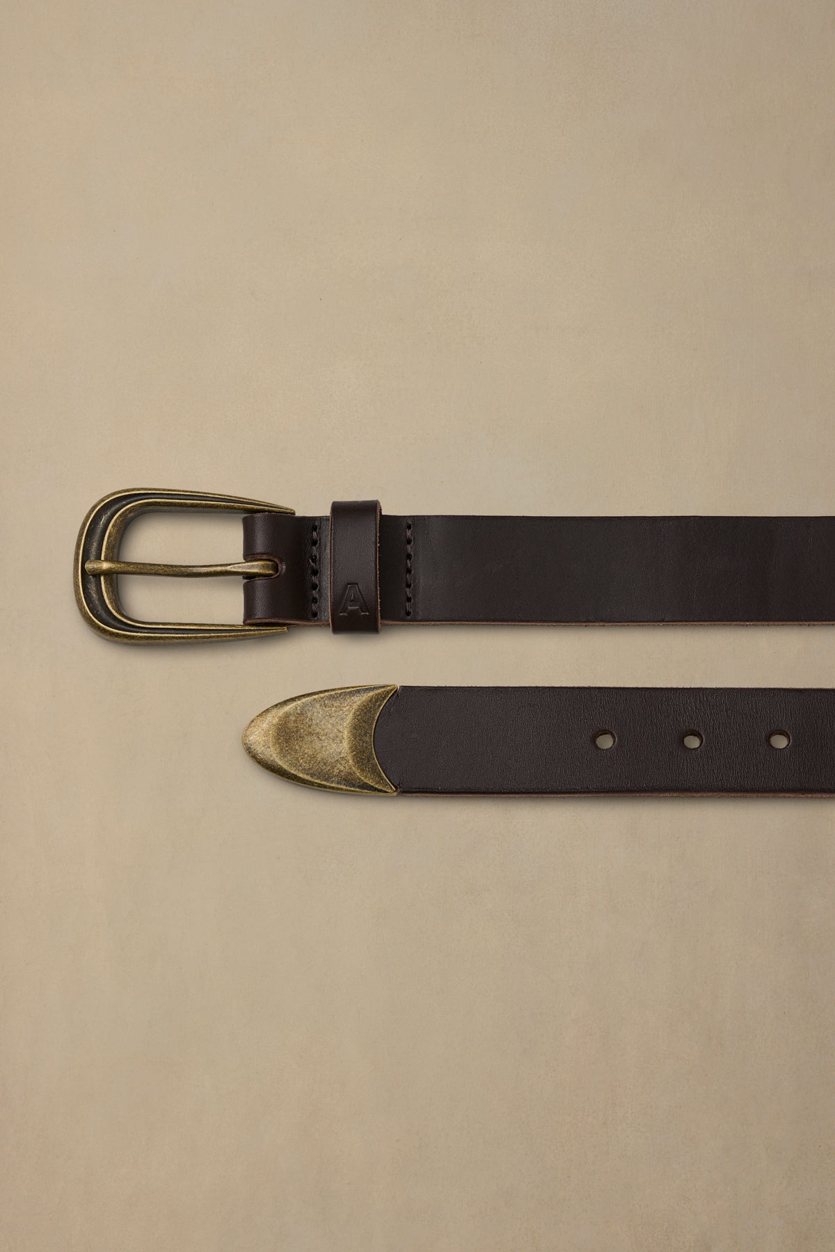 Dark brown leather belt with brass buckle and decorative tip, displayed flat on beige background