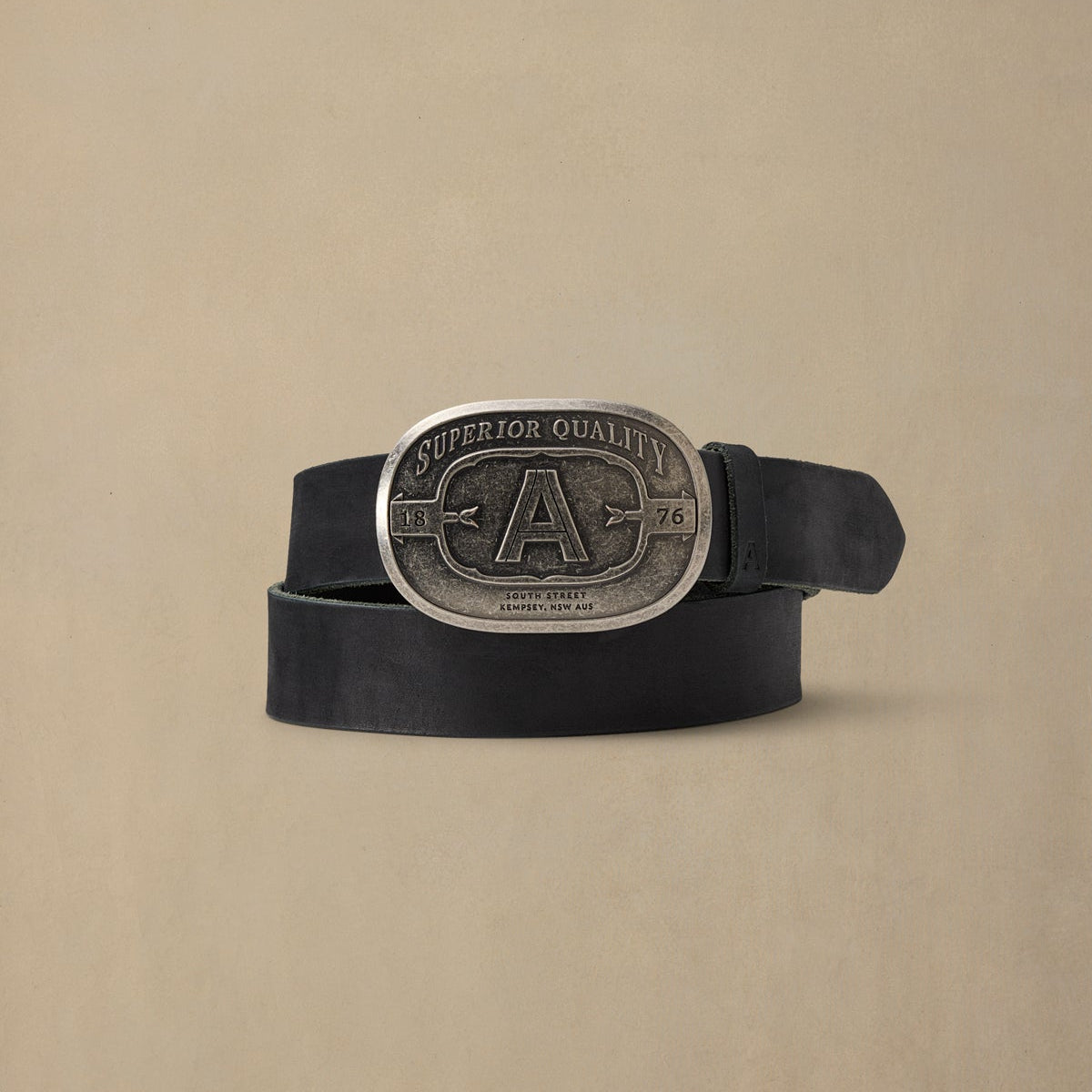 Black leather belt with silver pewter oval buckle featuring "A" initial and "Superior Quality" text, displayed on neutral beige background
