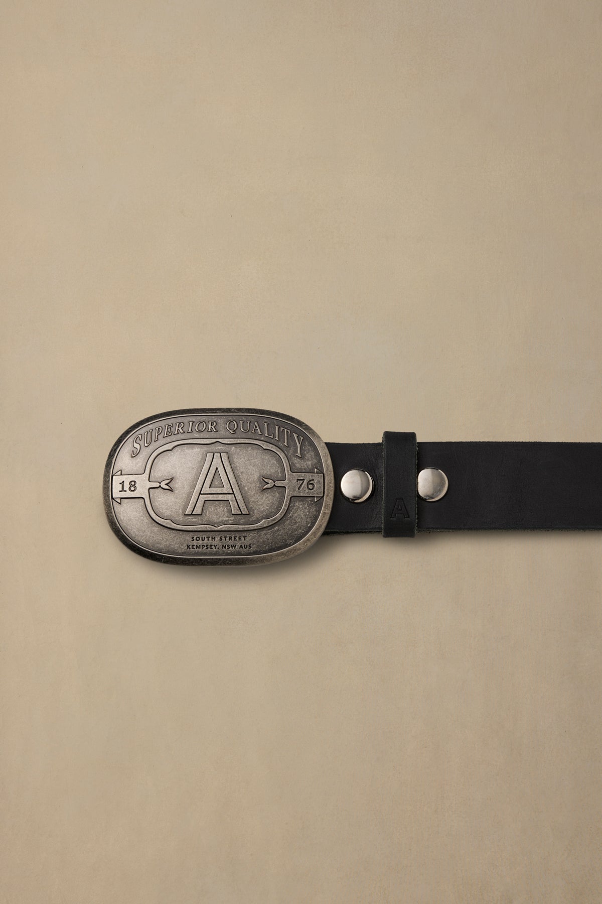 Pewter belt buckle with "A" logo and "Superior Quality" text on black leather strap against neutral background