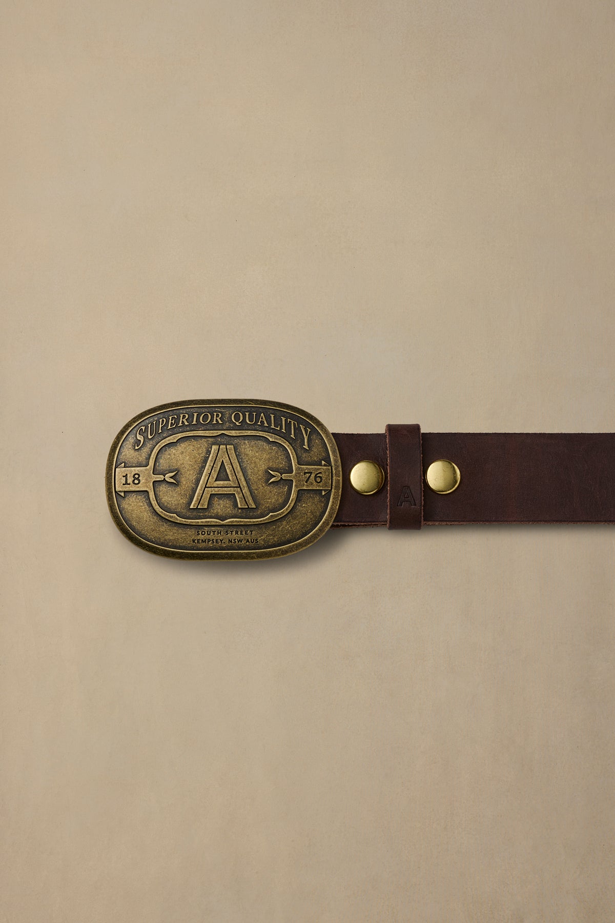 Chocolate brown leather belt with brass "A" buckle marked Superior Quality 1876, displayed flat against beige background