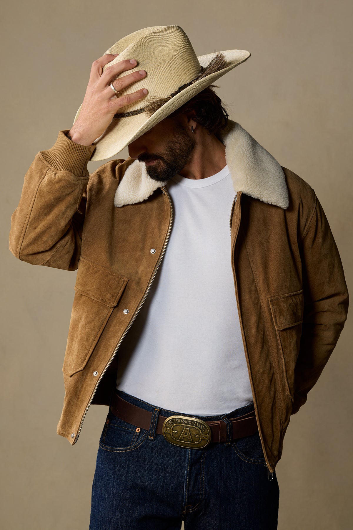 Chocolate brown leather belt with brass buckle worn with tan cowboy hat and sherpa-lined jacket on neutral background