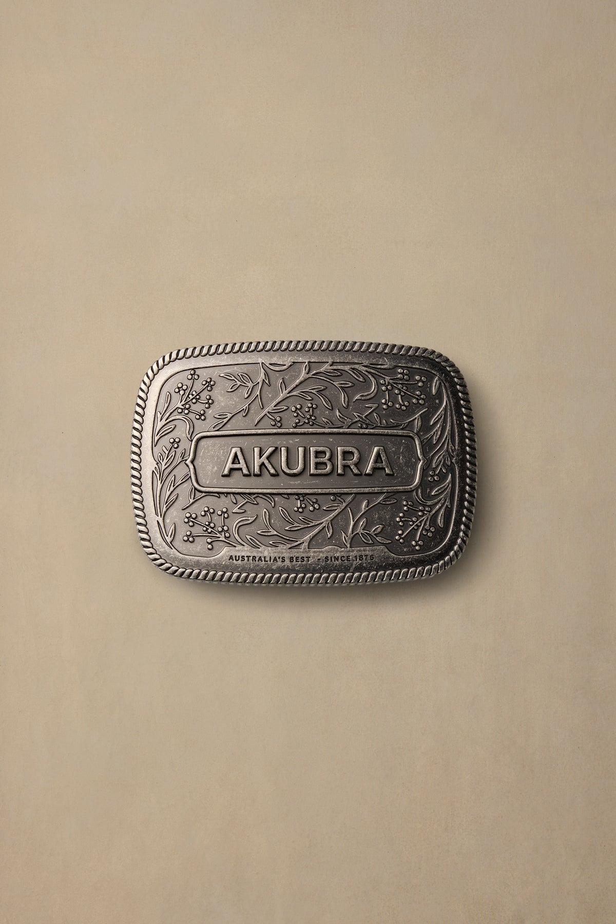 Silver pewter belt buckle with ornate floral engraving and rope border detail, displaying AKUBRA branding on beige background